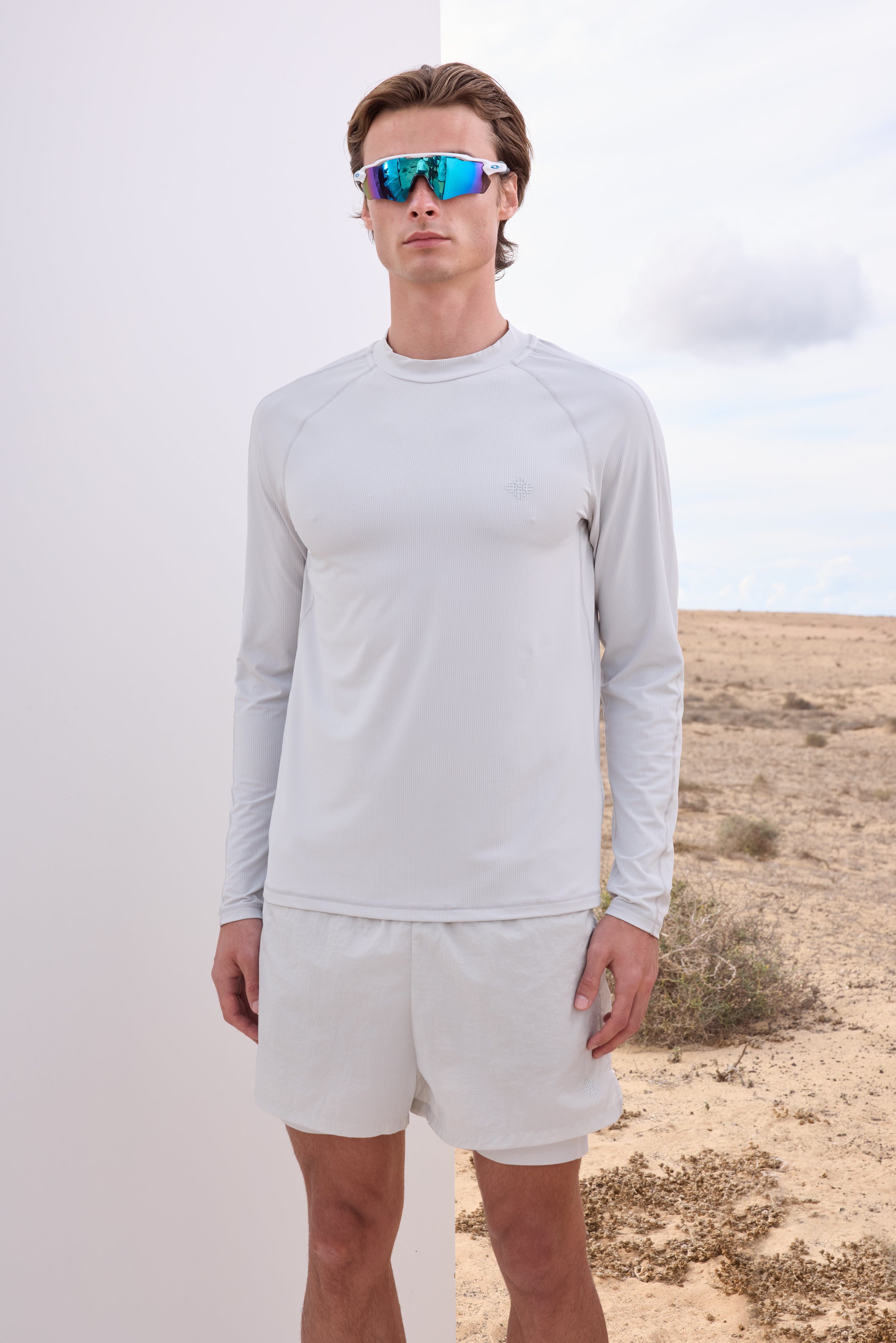 Man wearing a light grey long-sleeve t-shirt and white shorts with blue sunglasses in a desert setting.