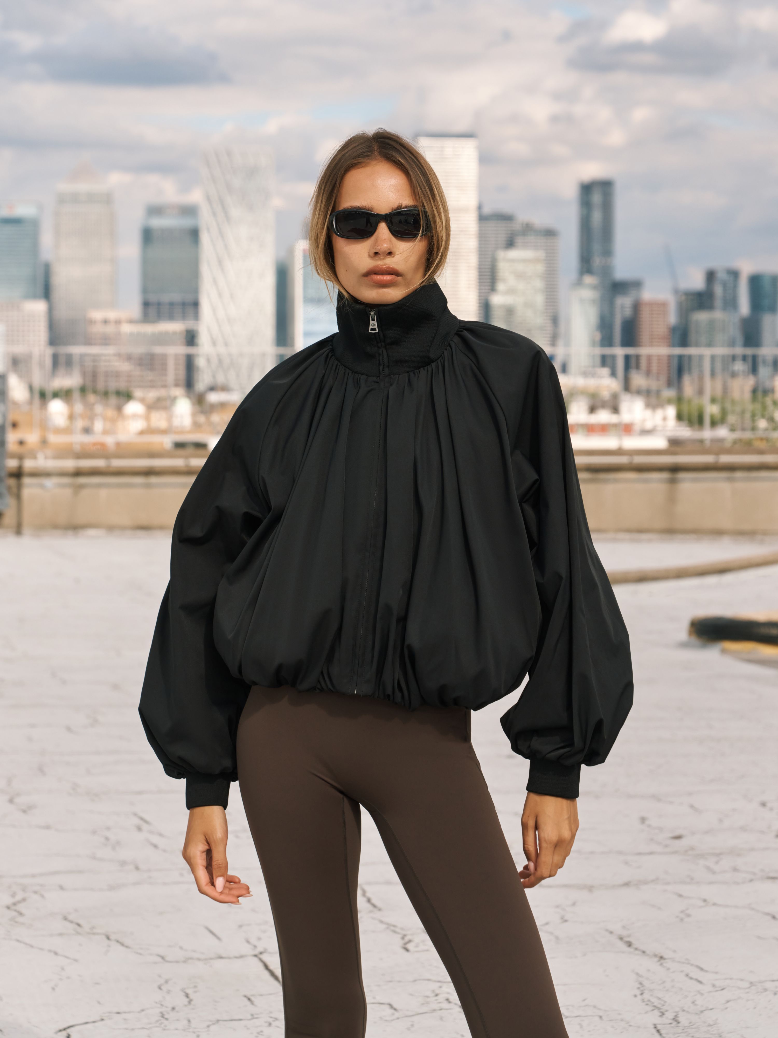 A model wearing a black high-neck, voluminous long-sleeve jacket with a gathered front and elastic cuffs, paired with dark brown leggings, standing against a city skyline background.