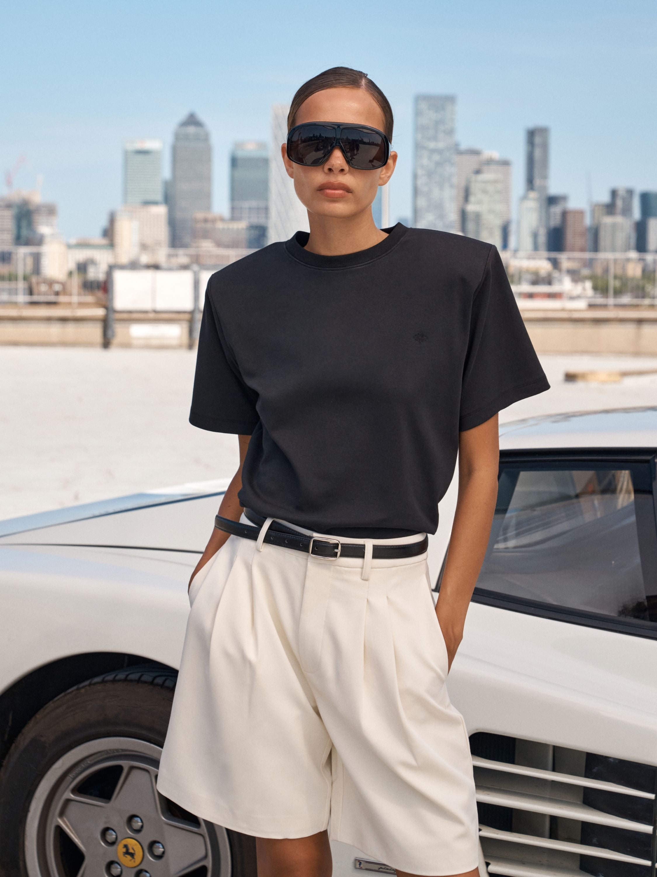 A model wearing a black short-sleeve t-shirt, off-white pleated shorts, a black belt, and black sunglasses stands in front of a white sports car with a city skyline in the background.