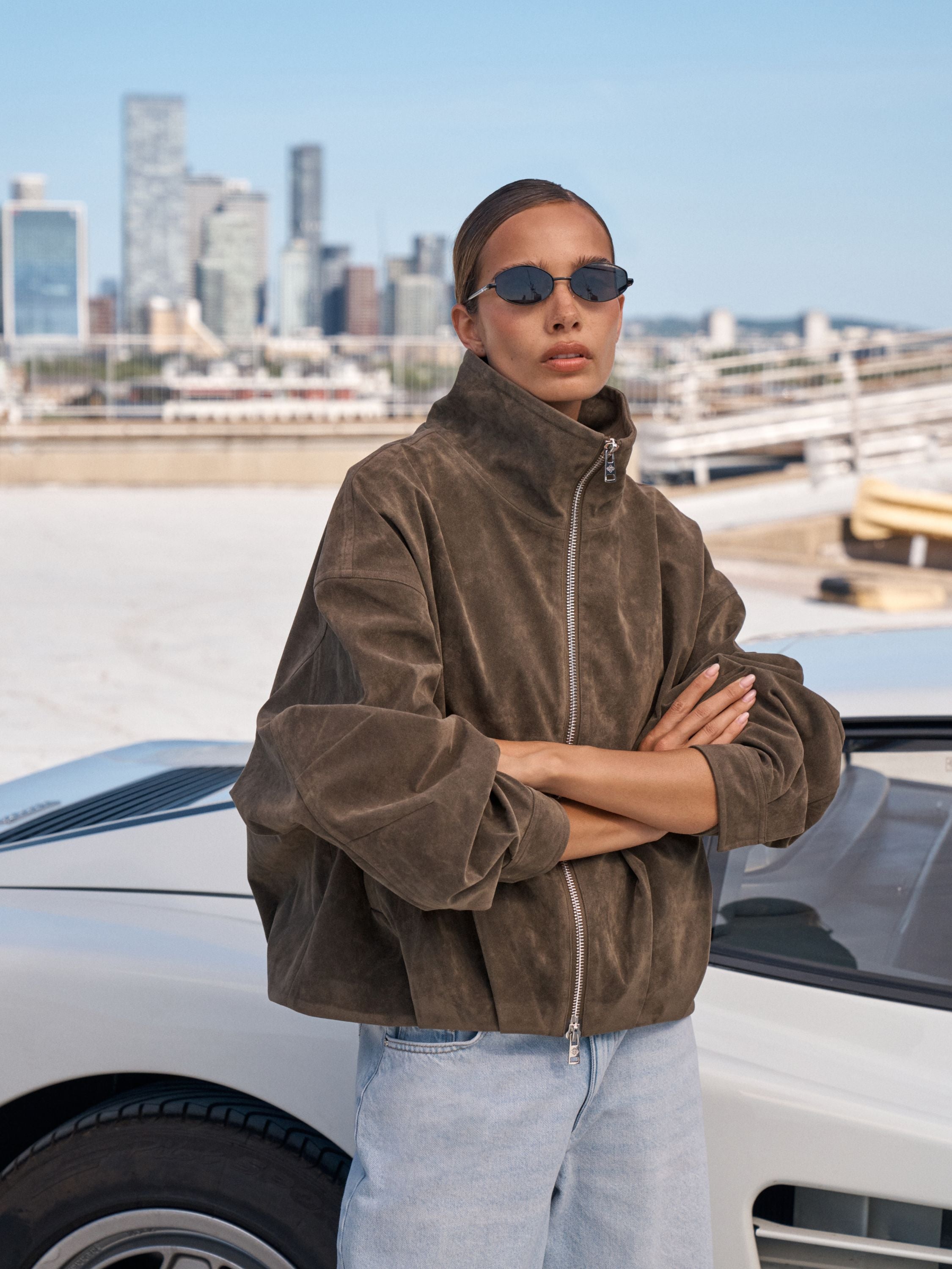 A model wearing a brown suede zip-up jacket and light blue jeans stands outdoors next to a white car with a city skyline in the background.