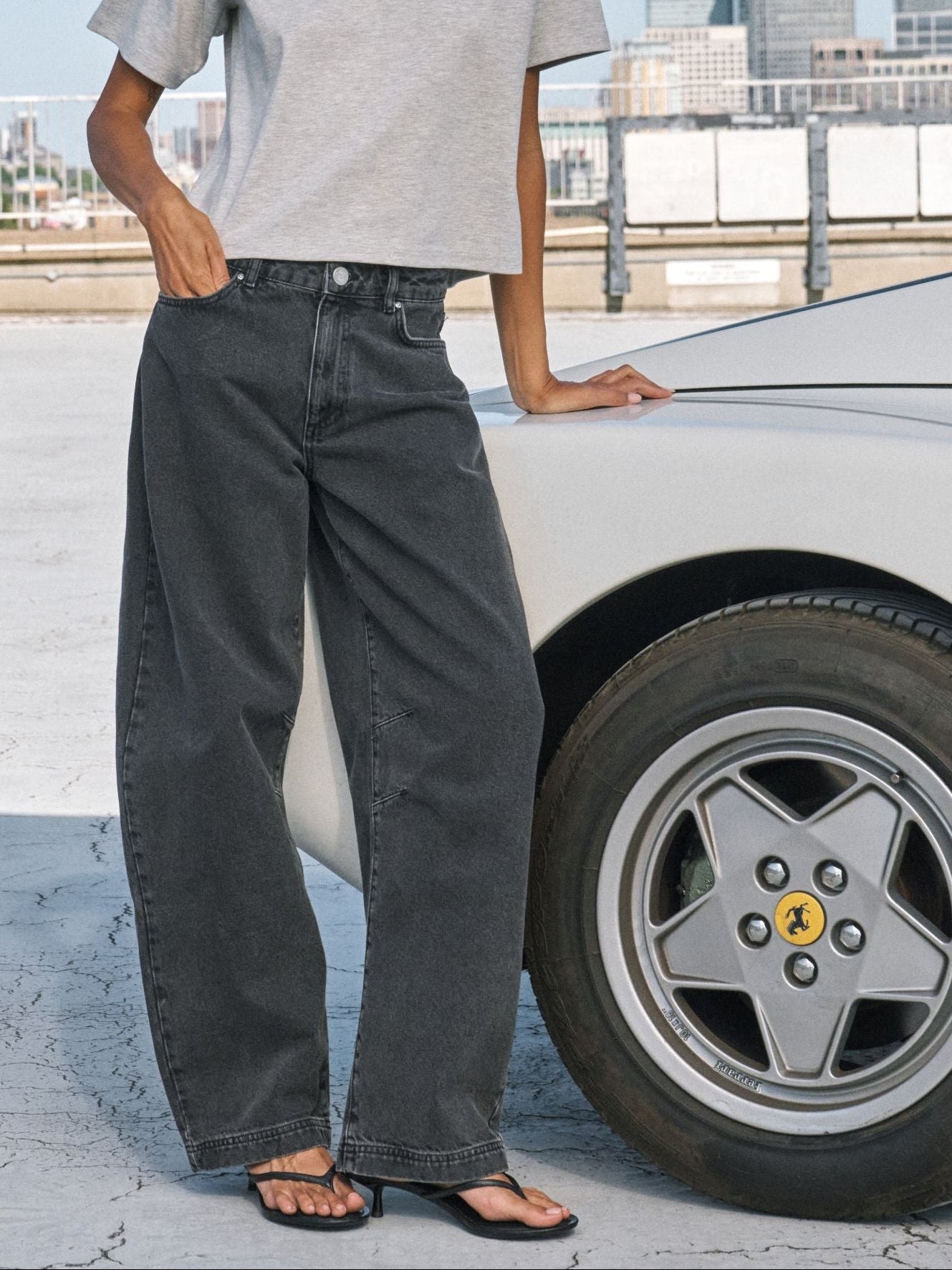 A person wearing black wide-leg denim jeans, a light gray t-shirt, and black sandals stands next to a white car, with a Ferrari wheel visible.