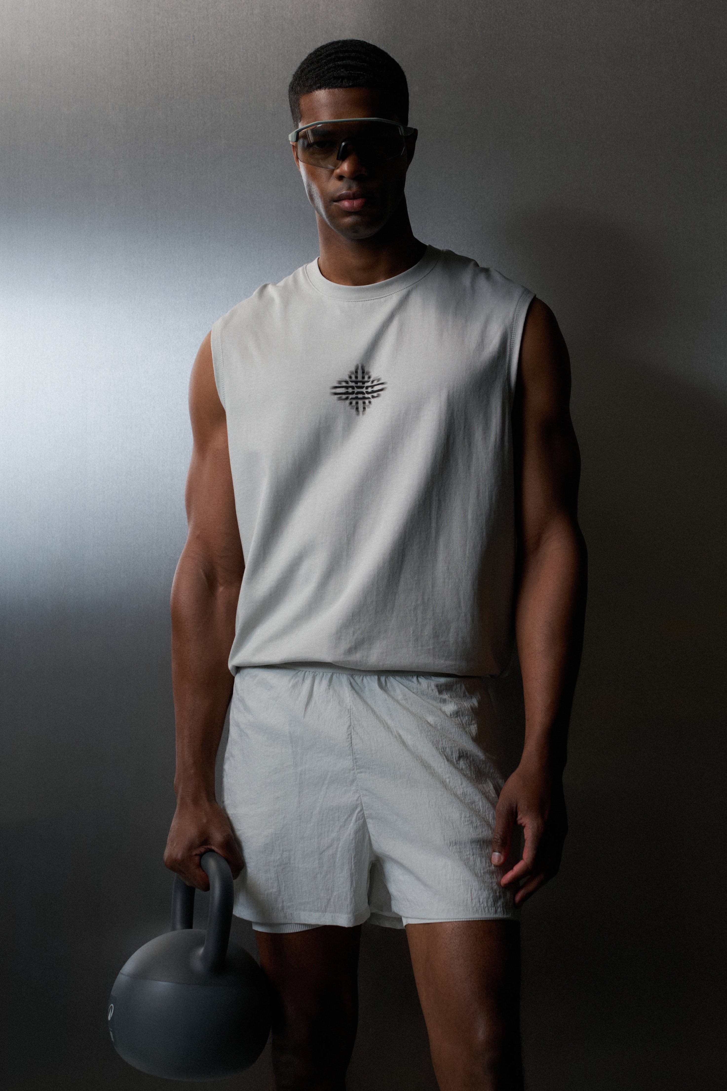 Man wearing a white sleeveless shirt and shorts holding a kettlebell against a dark background
