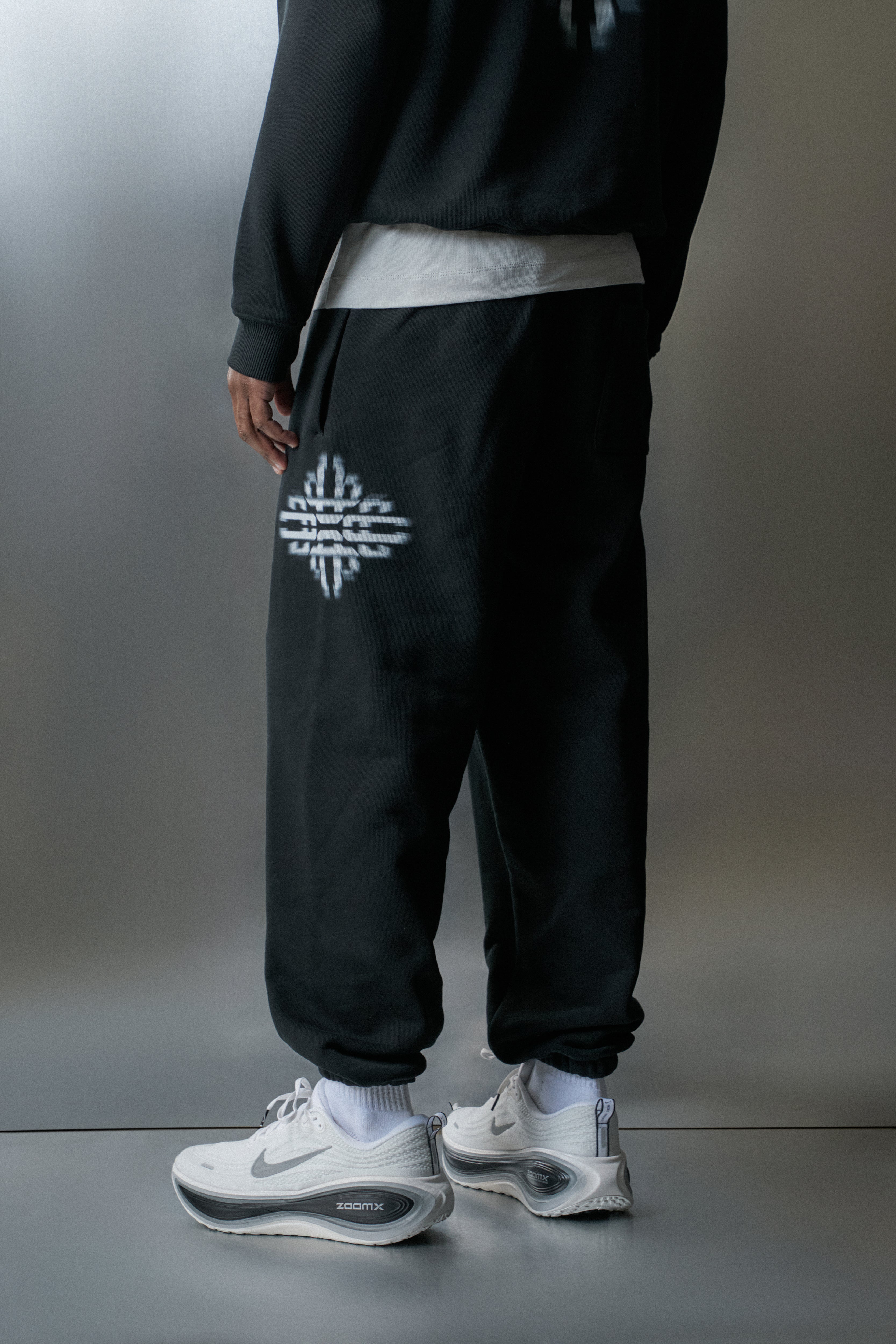Person wearing black jogger pants with a logo and white sneakers on a grey background