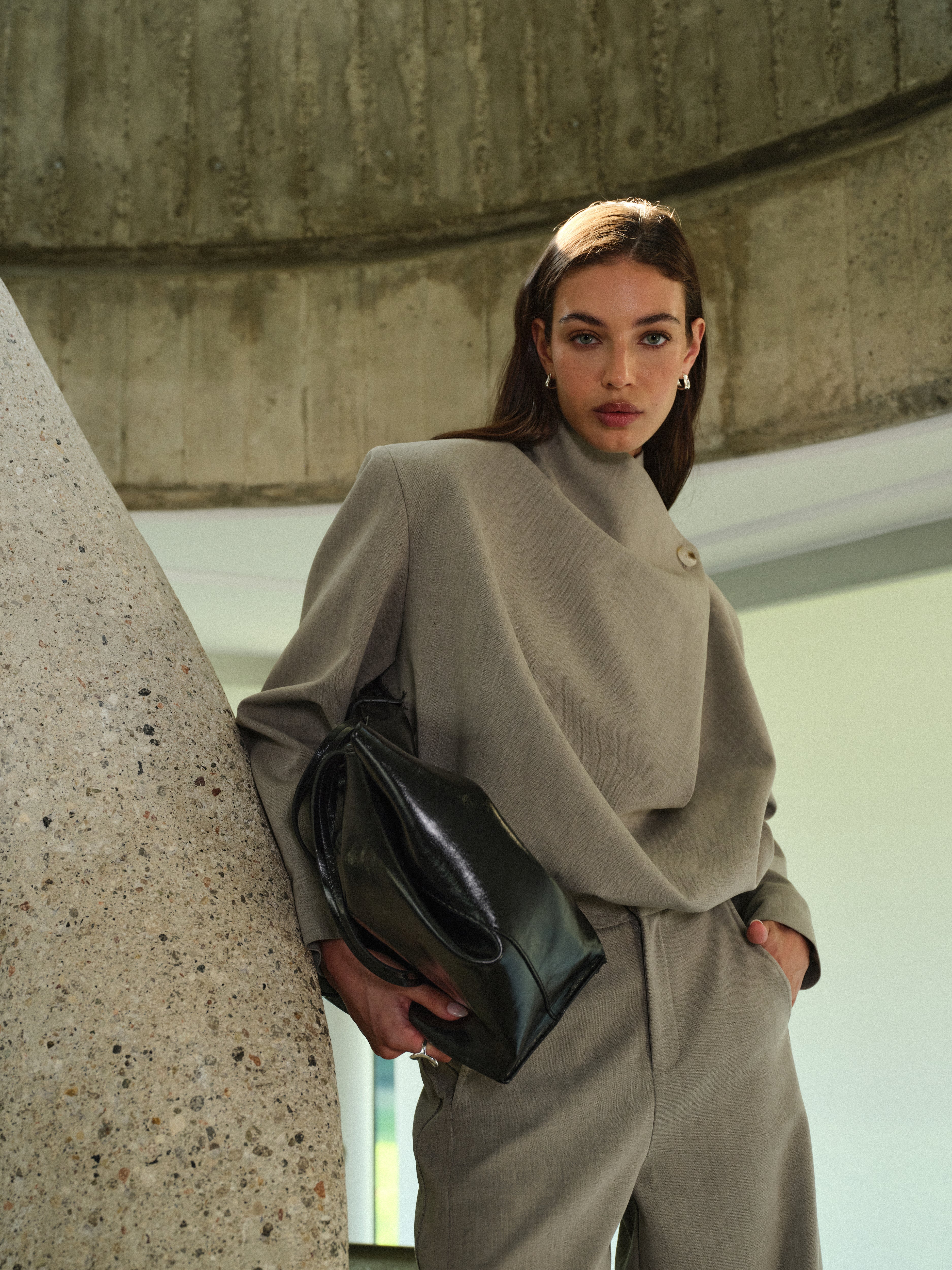 Woman in a grey tailored set holding a black bag against a concrete wall.