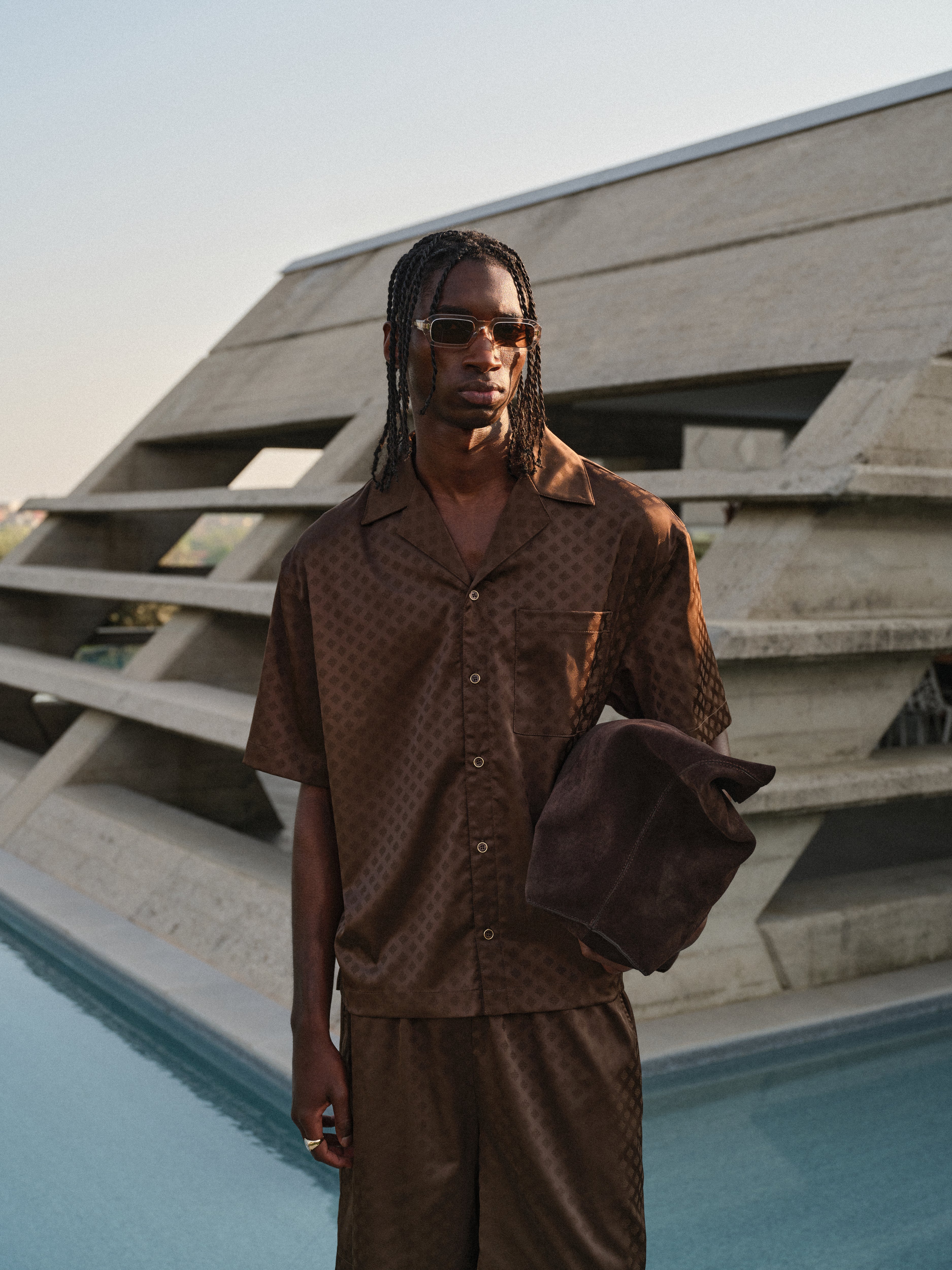 Man in a brown satin shirt and matching satin shorts standing by a pool with a modern architectural background