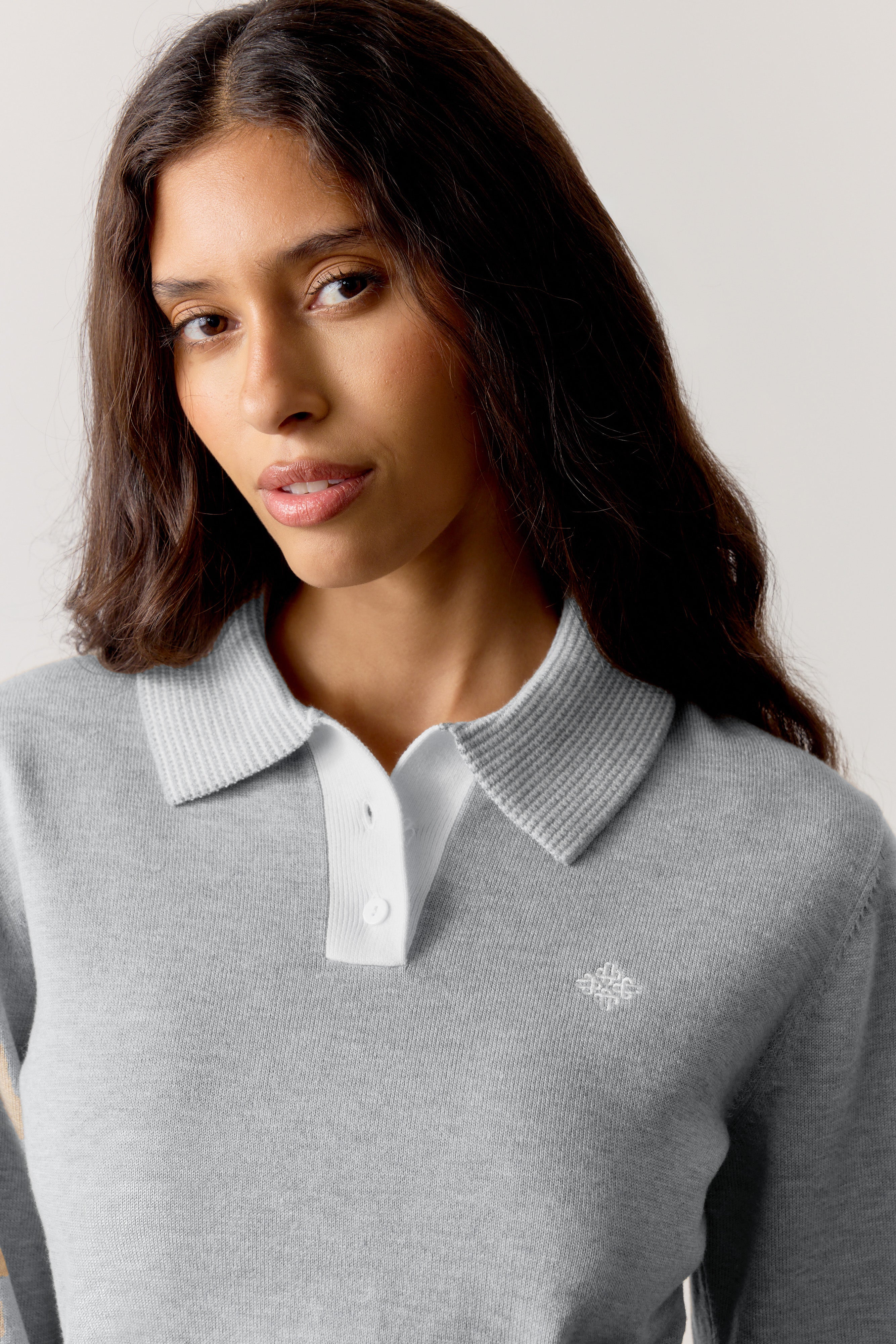 Woman wearing a grey long sleeved polo with a white collar and logo on a light background.
