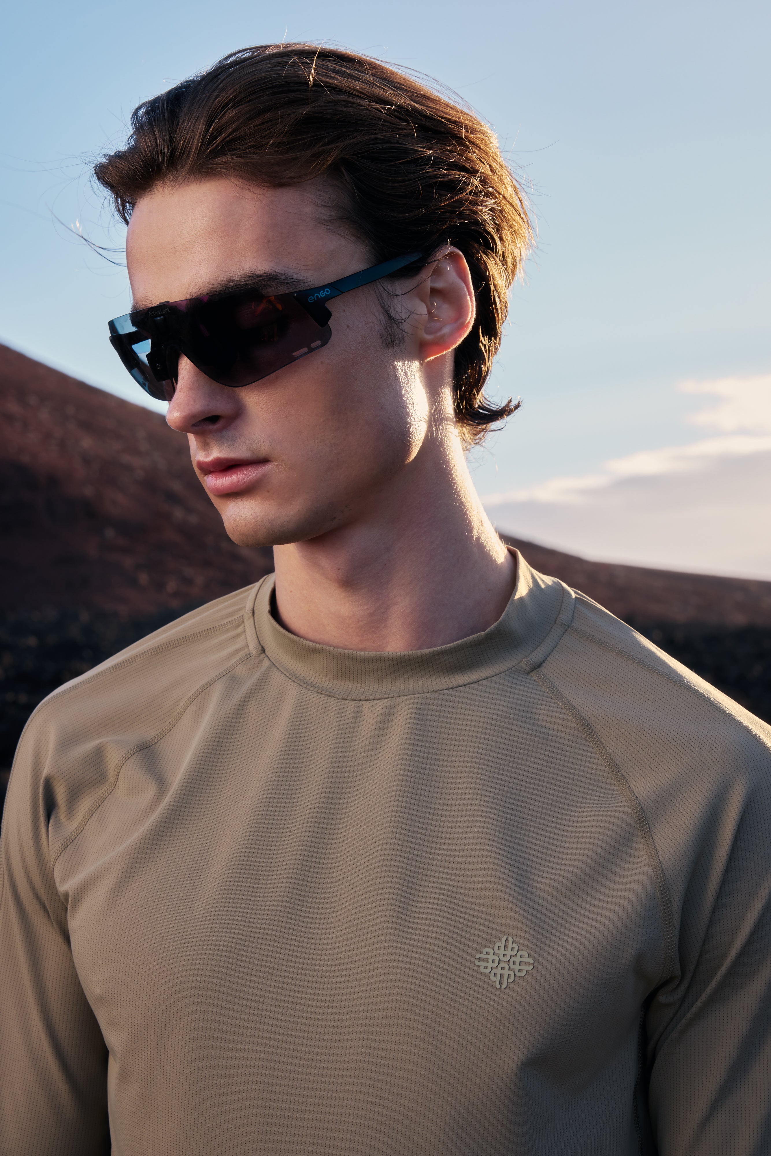 Man wearing sunglasses and a khaki long-sleeve shirt with a logo against a mountainous background