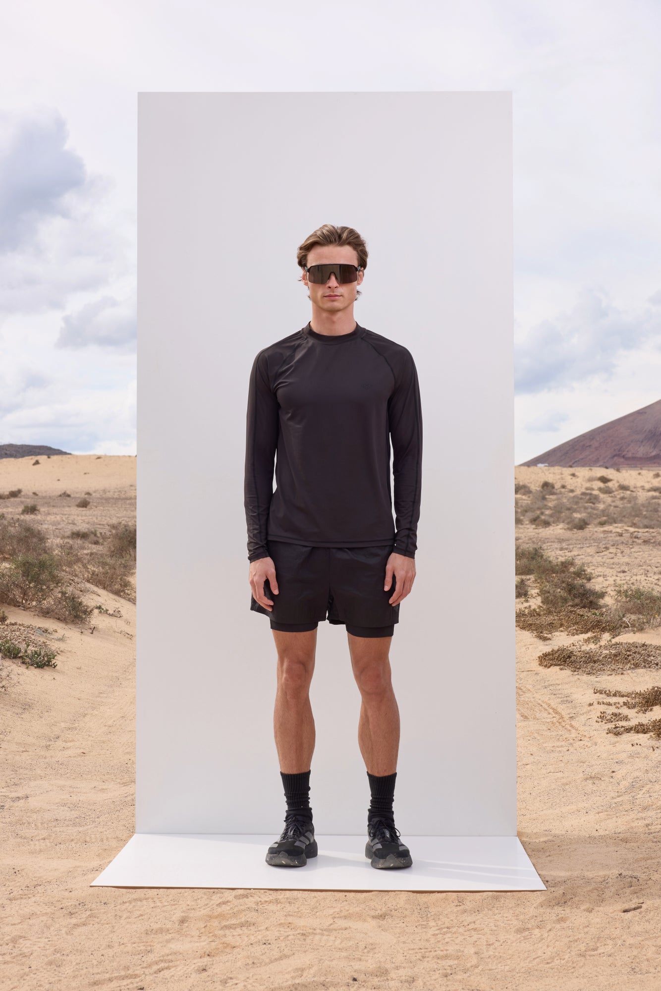 Person wearing a black outfit standing in front of a white panel with a desert landscape in the background