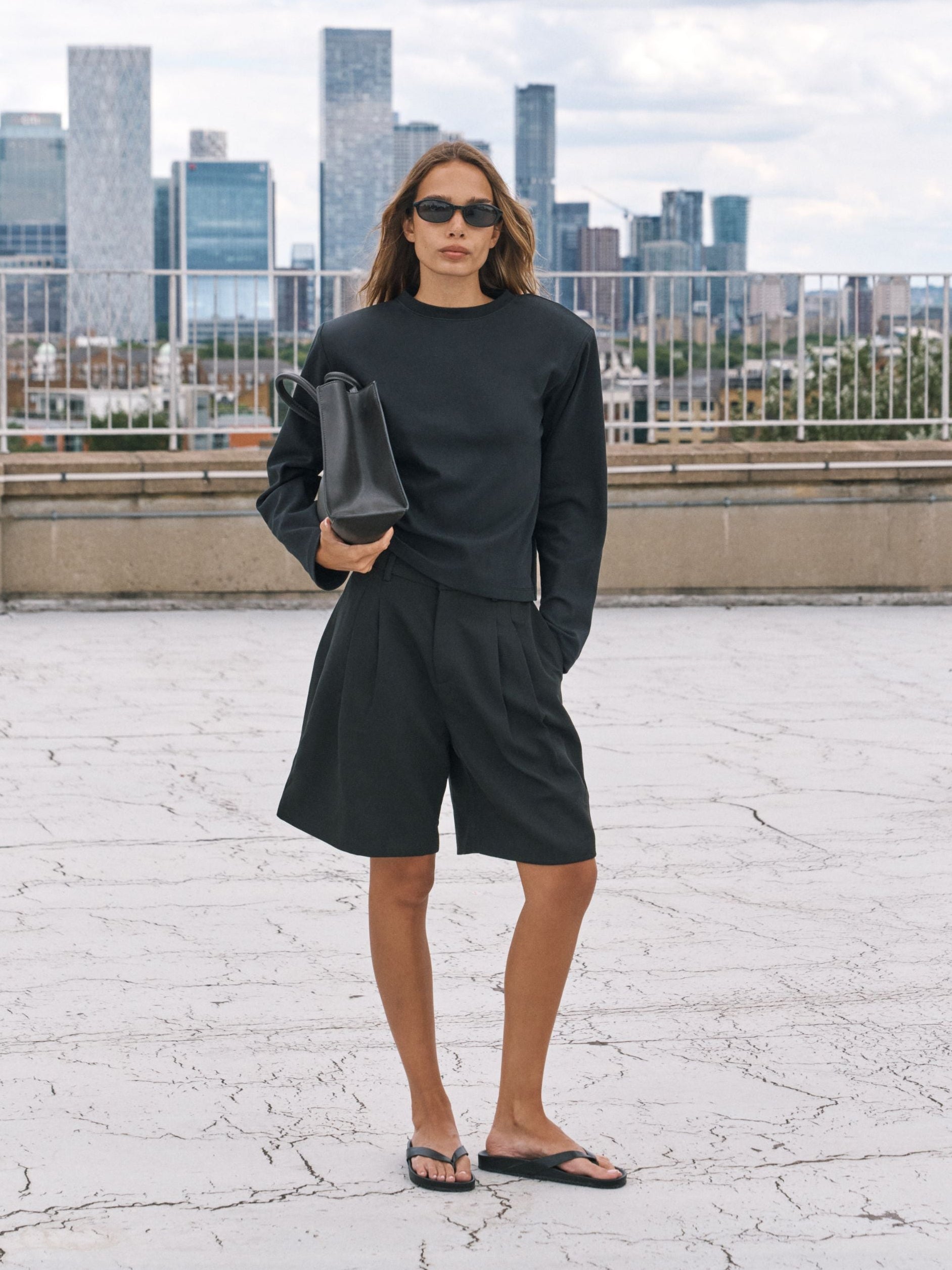 A woman stands on a rooftop wearing a matching black long-sleeve top and shorts set, black sunglasses, black sandals, and holding a black handbag, with a city skyline in the background.