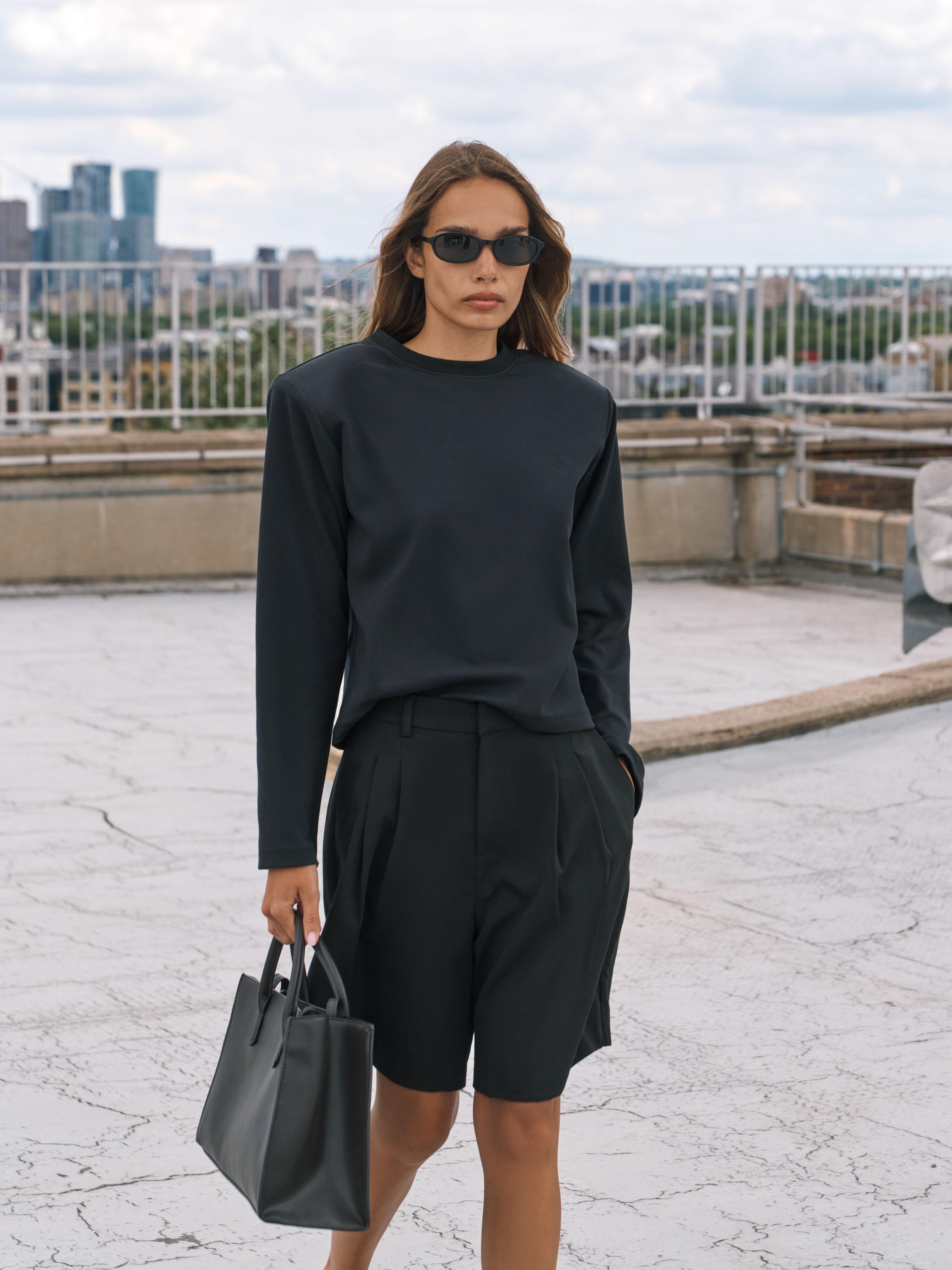 A woman stands on a rooftop terrace wearing a black long-sleeve top and black shorts, holding a black tote bag. A city skyline is visible in the background under a cloudy sky.
