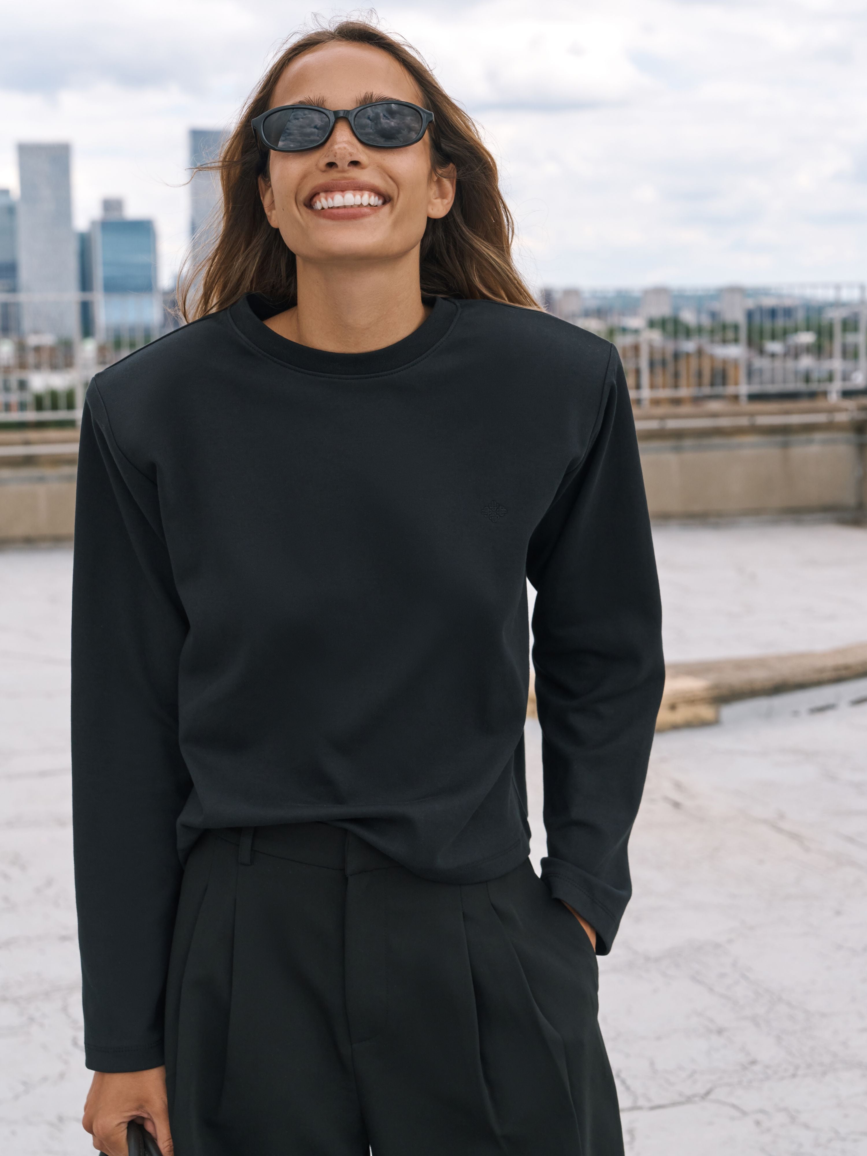 A woman wearing a black long-sleeve crewneck top and black trousers, standing on a rooftop with a city skyline in the background under an overcast sky.