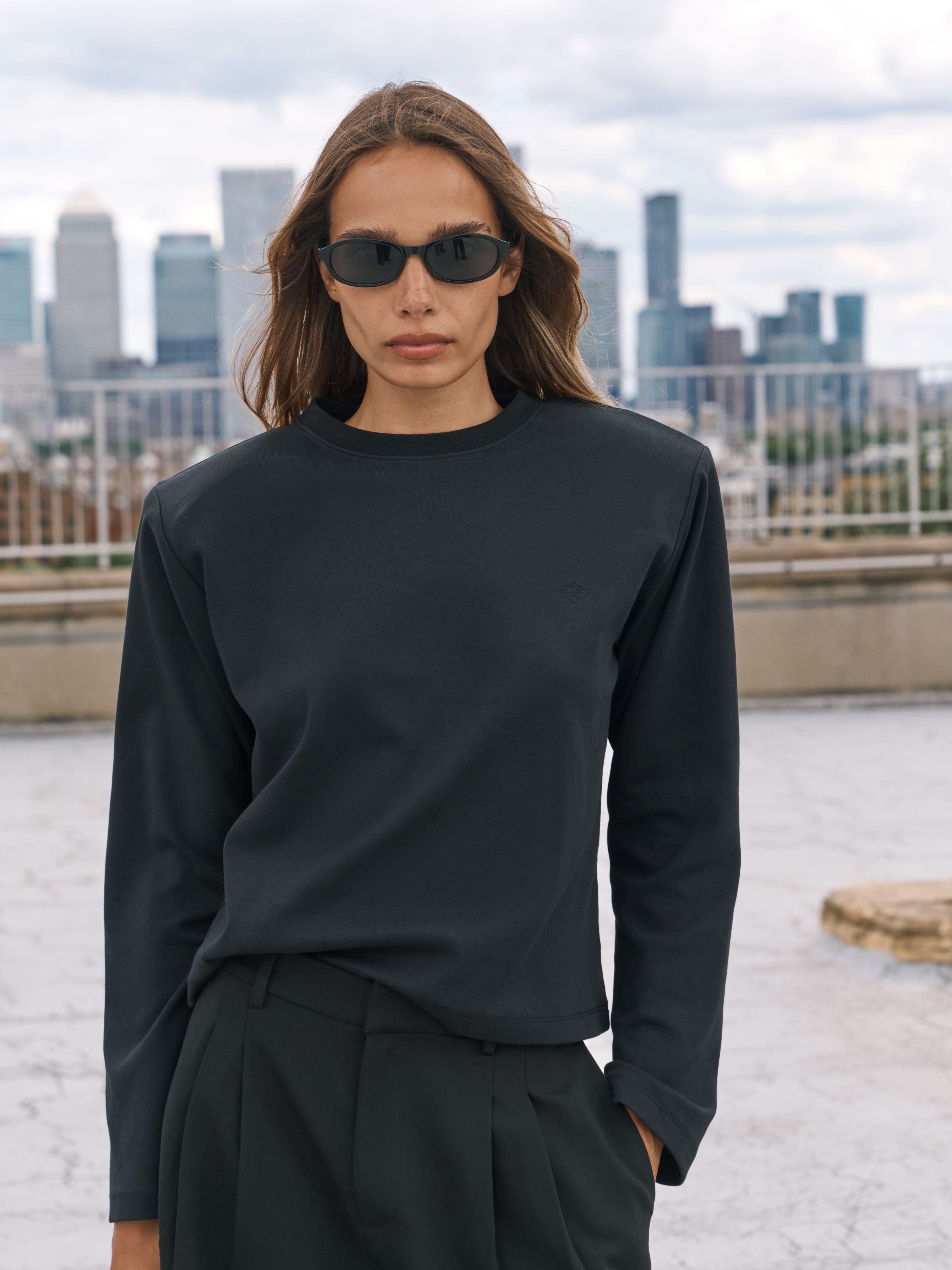 A woman wearing a black long-sleeve t-shirt and black pants, standing on a rooftop with a city skyline in the background.