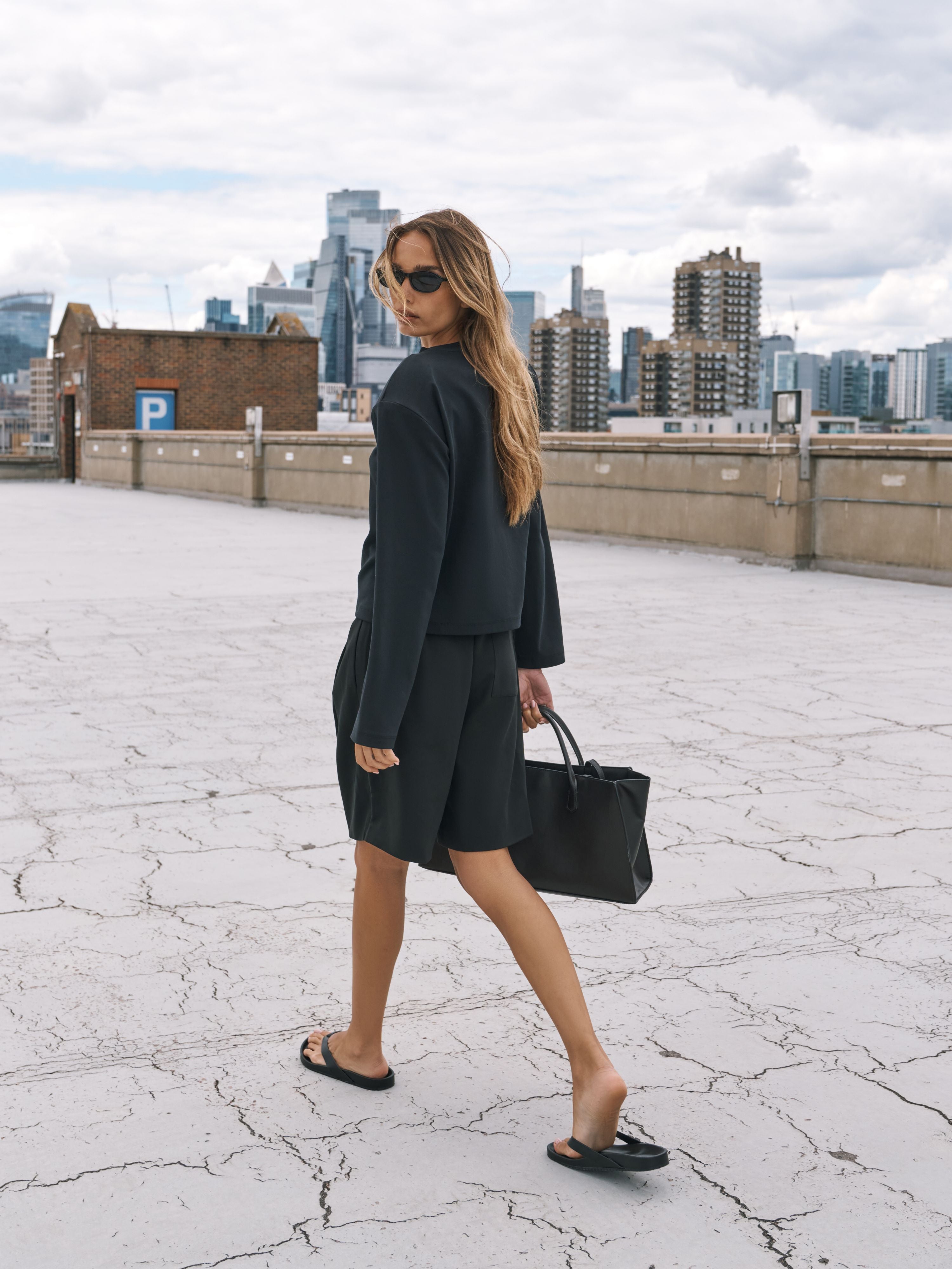 A woman wearing a long sleeved black t-shirt and pleated shorts walks on a cracked concrete rooftop with a city skyline in the background, carrying a black tote bag.