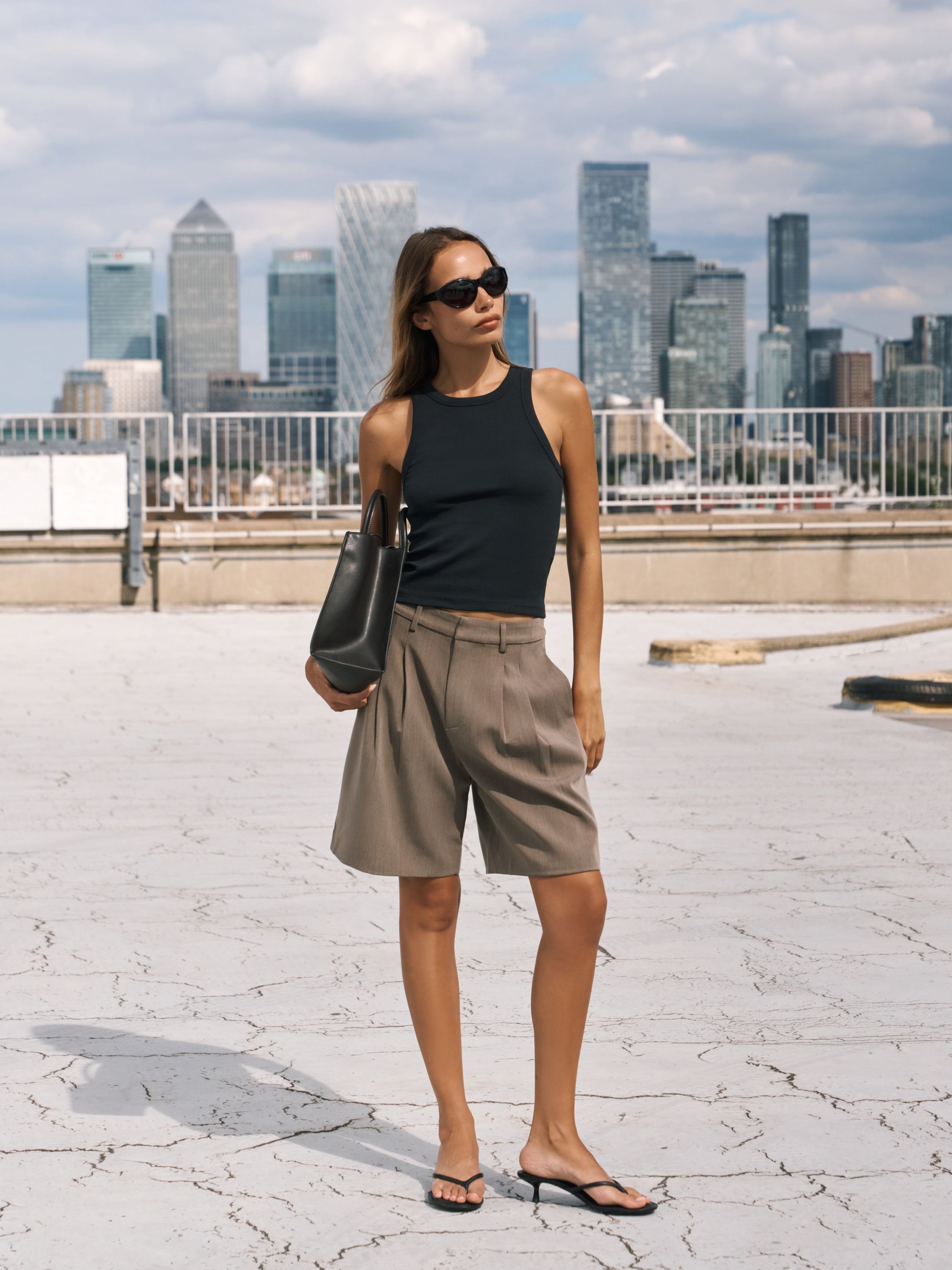 A woman stands on a rooftop wearing a black sleeveless top, high-waisted beige shorts, black sandals, and sunglasses, with a city skyline in the background.