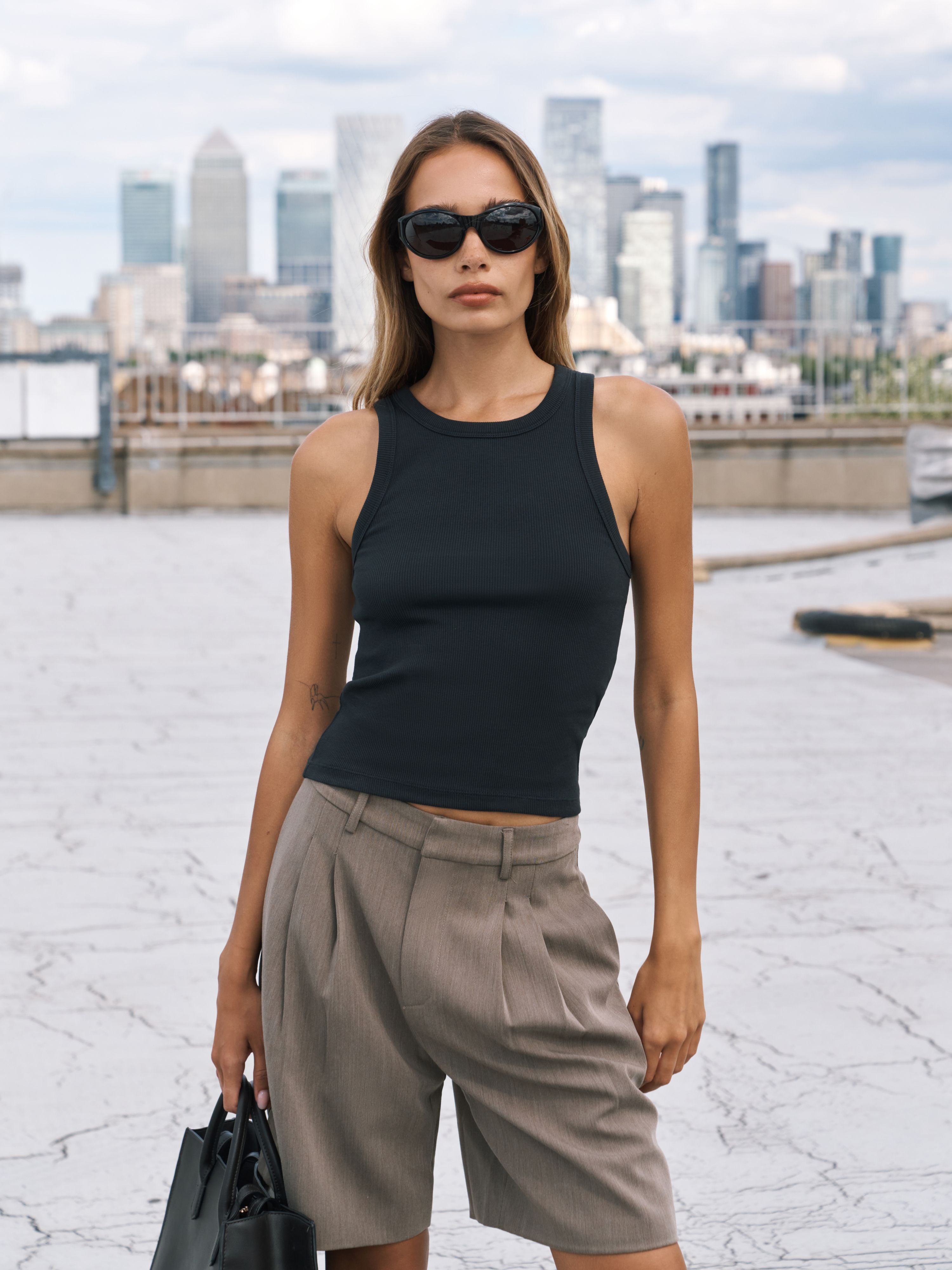 A woman stands on a rooftop wearing a fitted black vest and taupe pleated shorts, with a city skyline in the background. She is also wearing dark sunglasses and holding a black handbag.
