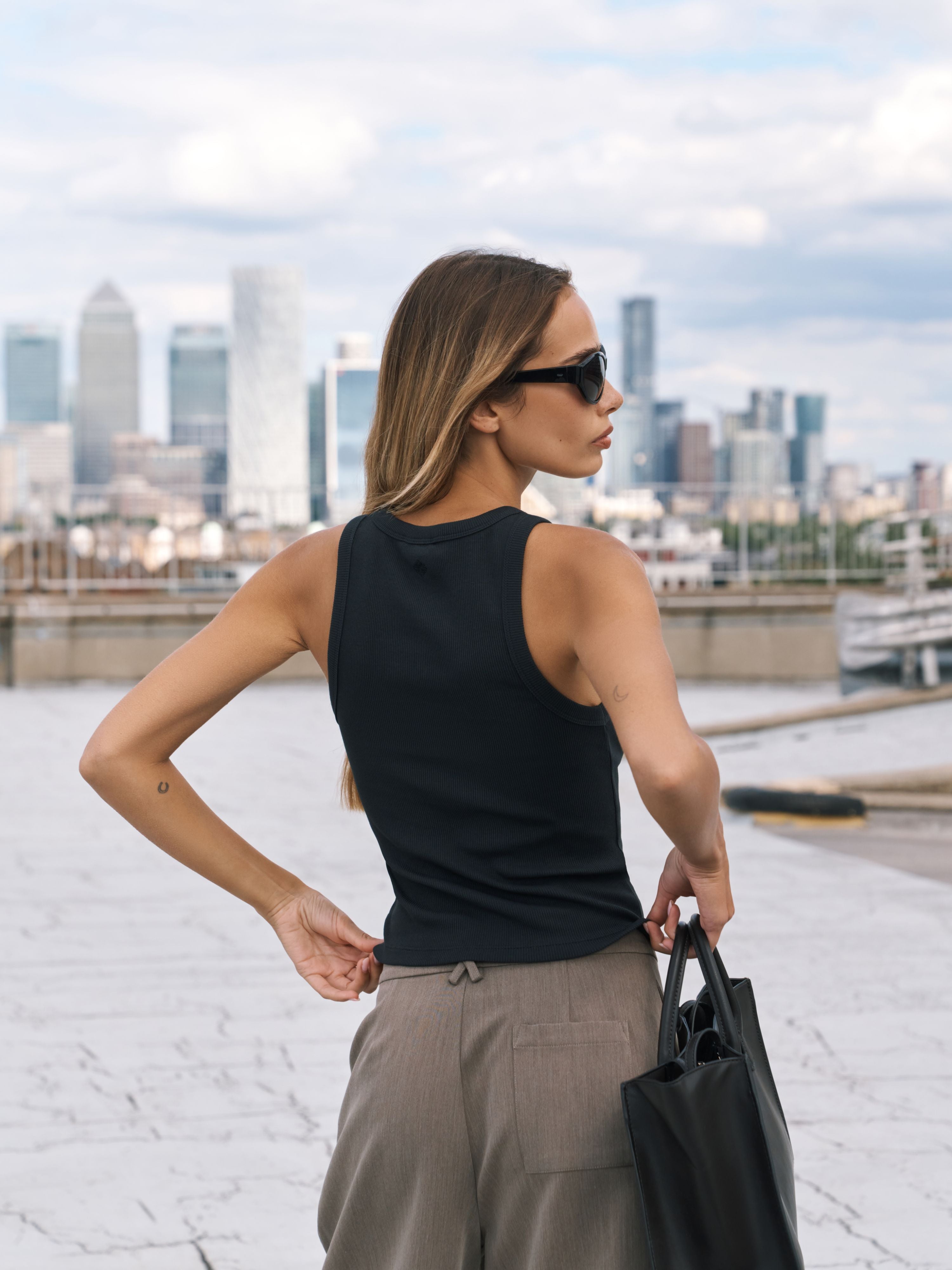 A woman stands on a rooftop wearing a black sleeveless tank top and light brown wide-leg trousers, holding a black tote bag, with a city skyline in the background.