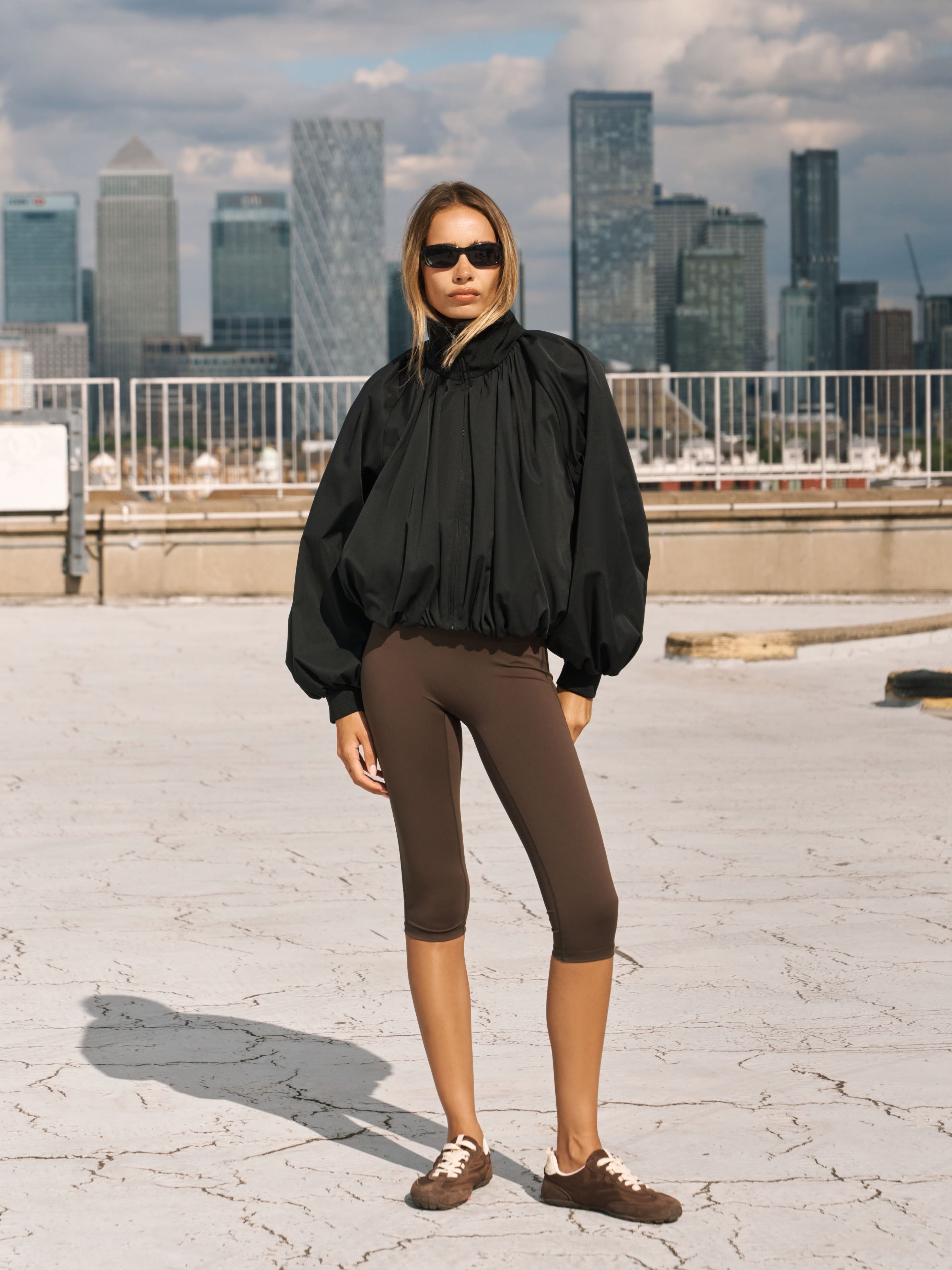A woman stands on a rooftop wearing a black oversized long-sleeve blouse with dramatic puffed sleeves, brown capri leggings, and brown sneakers, with a city skyline in the background.