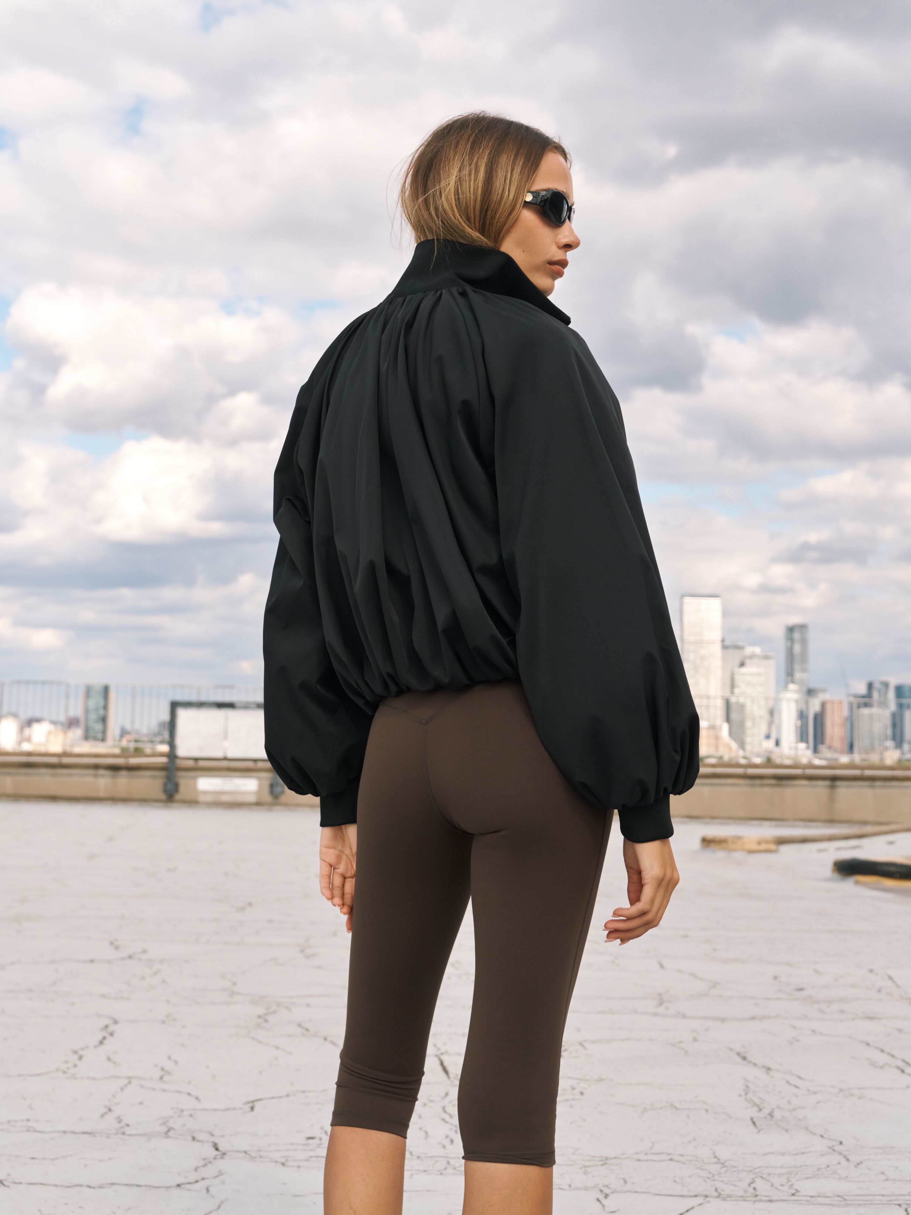 A woman wearing a black oversized balloon-sleeve jacket and brown capri leggings, photographed from behind on a rooftop with a cloudy sky and city skyline in the background.