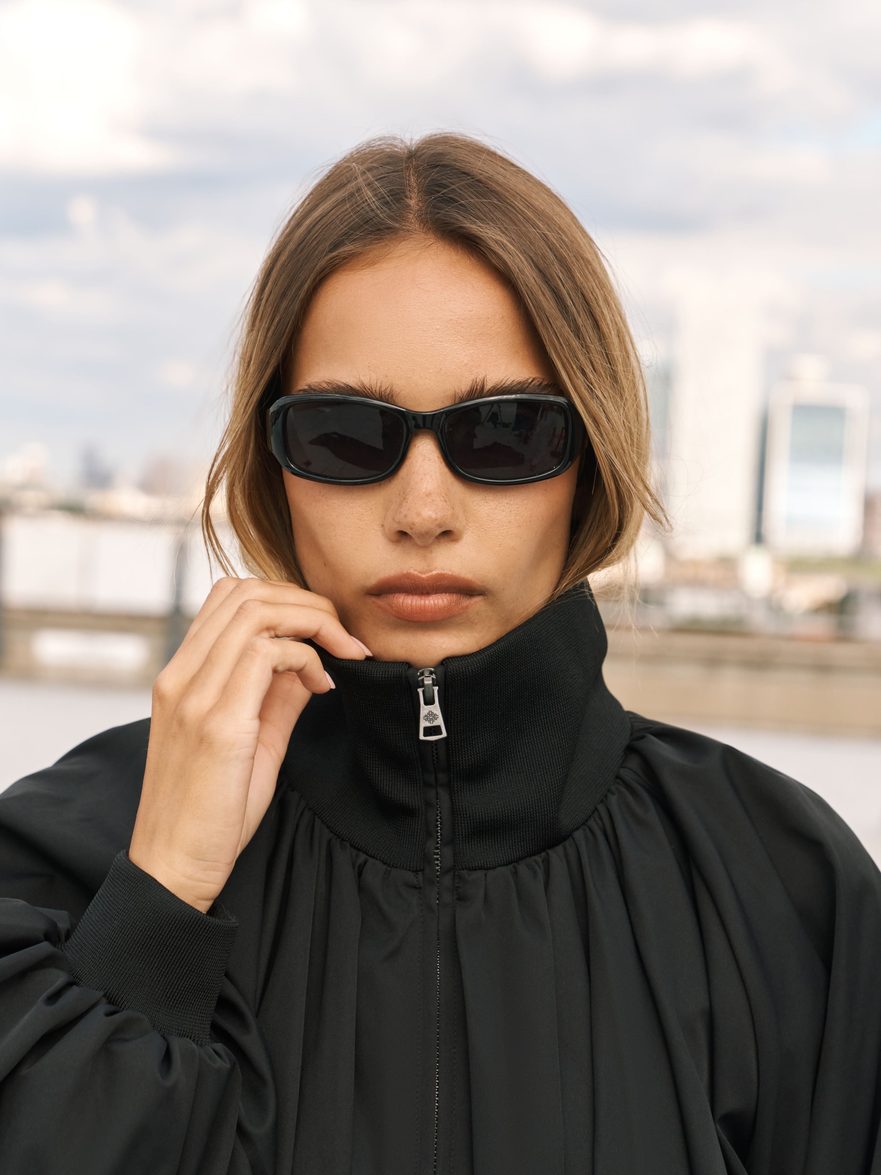 A woman wearing black cat-eye sunglasses and a black high-collar zip-up jacket, posing outdoors with a blurred city skyline and river in the background.
