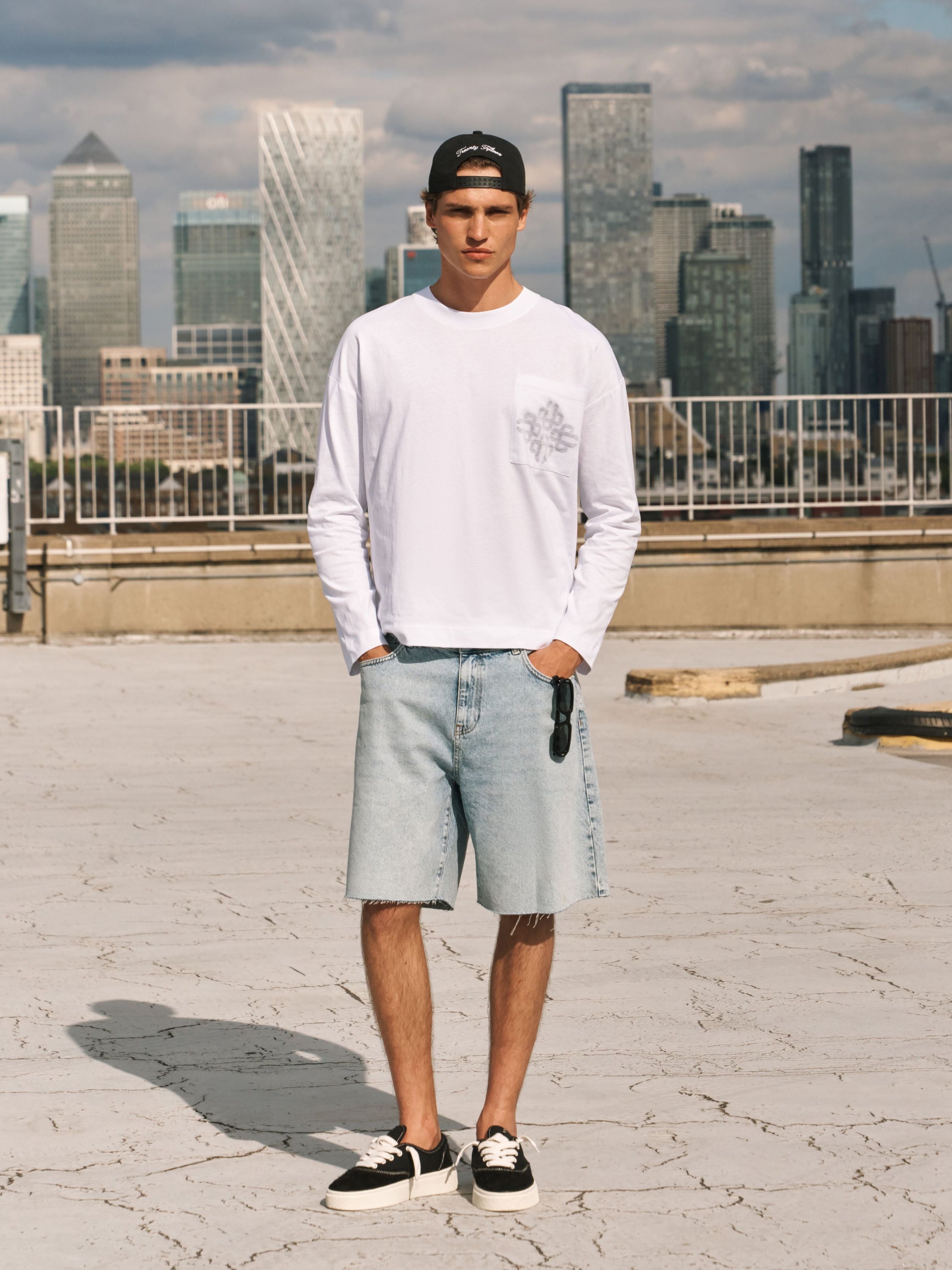 A man stands on a rooftop wearing a white long-sleeve crewneck shirt, light-wash denim shorts, black and white sneakers, and a black baseball cap, with a city skyline in the background.