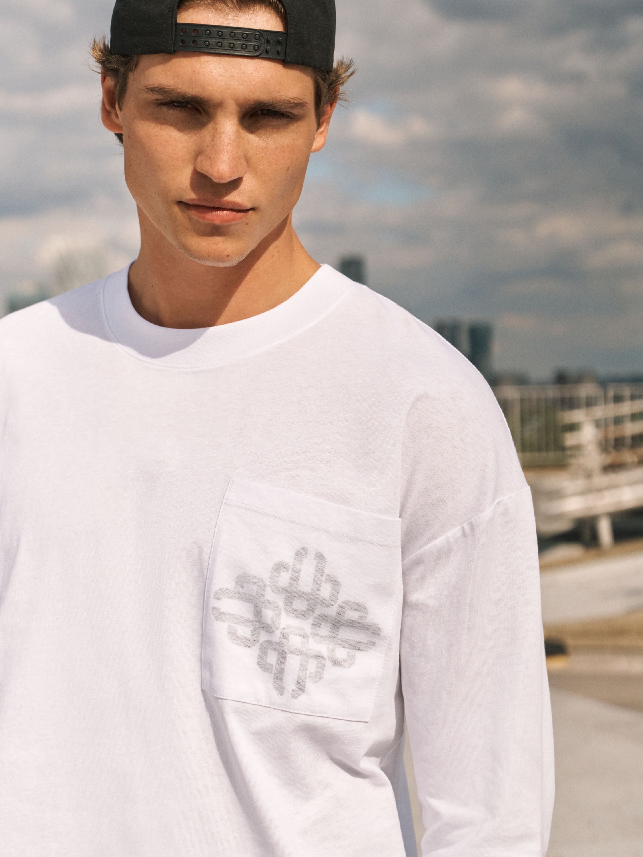 A man wearing a white long-sleeve pocket T-shirt with a gray graphic print on the chest pocket, paired with a black baseball cap, set against an outdoor urban background.