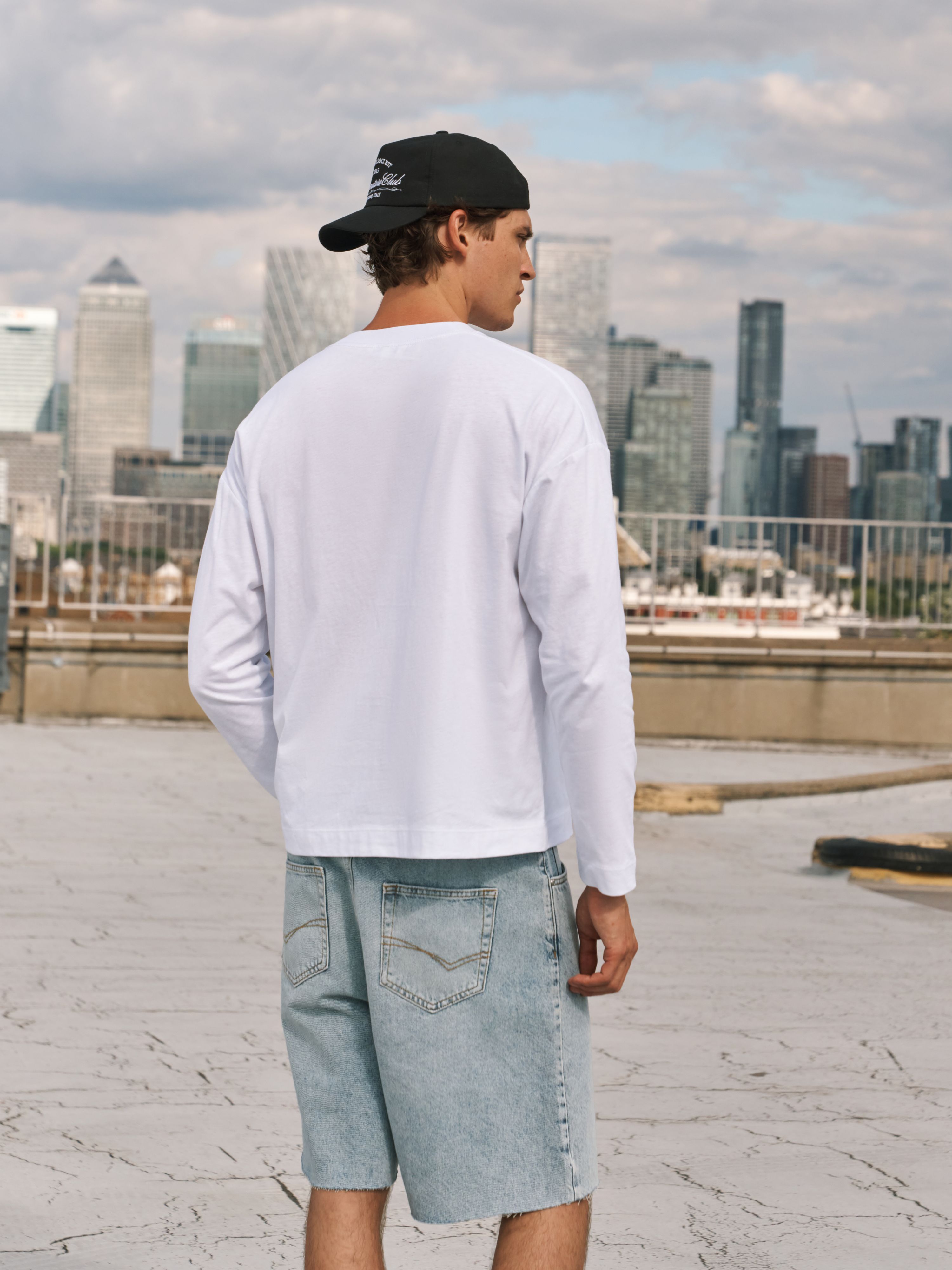A man wearing a white long-sleeve t-shirt, light wash denim shorts, and a black baseball cap, standing on a rooftop with a city skyline in the background.