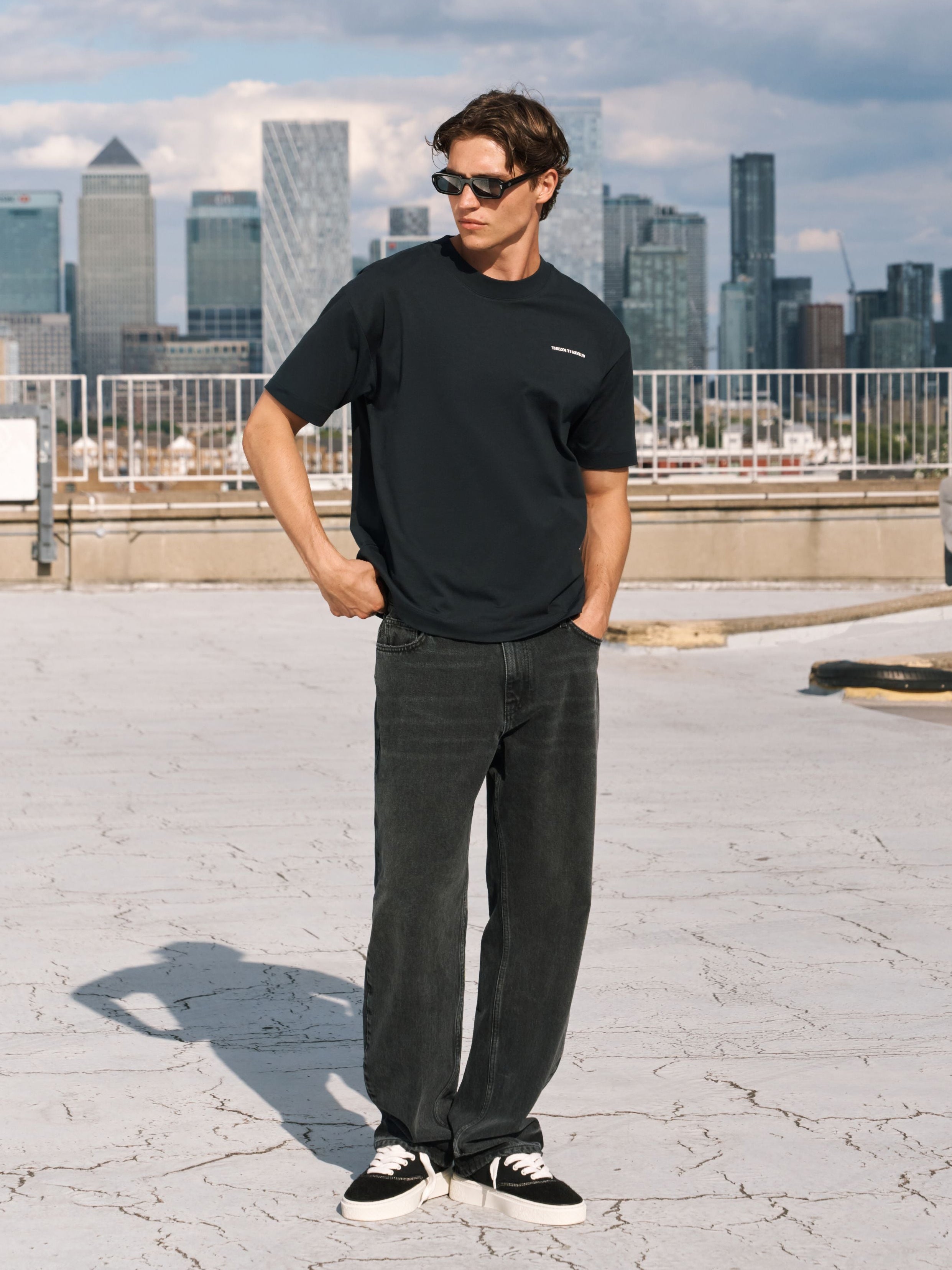 A man wearing a black crewneck t-shirt with white graphic prints on the sleeves, dark gray jeans, and black-and-white sneakers, standing on a rooftop with a city skyline in the background.