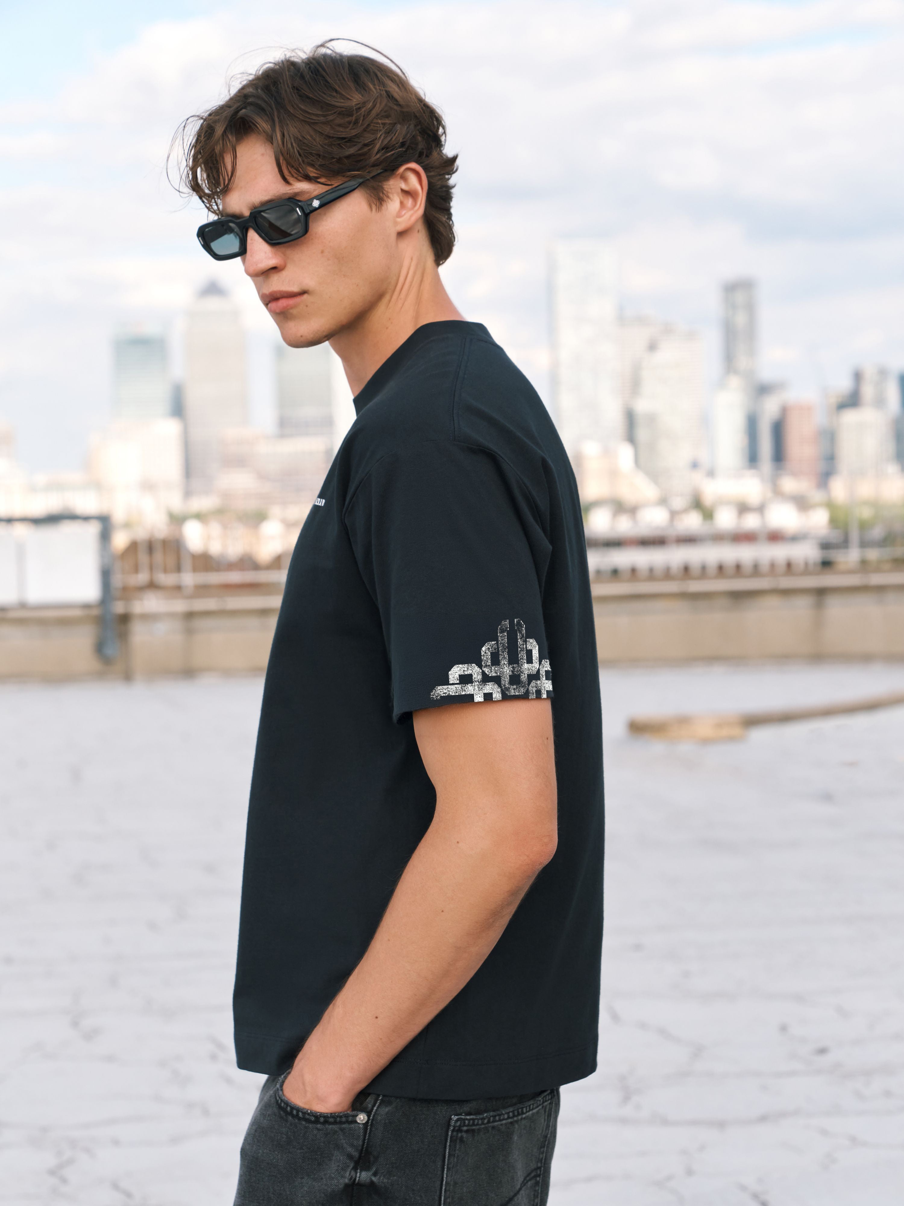 A man wearing a black short-sleeve t-shirt with a white graphic print on the sleeve, standing on a rooftop with a city skyline in the background.