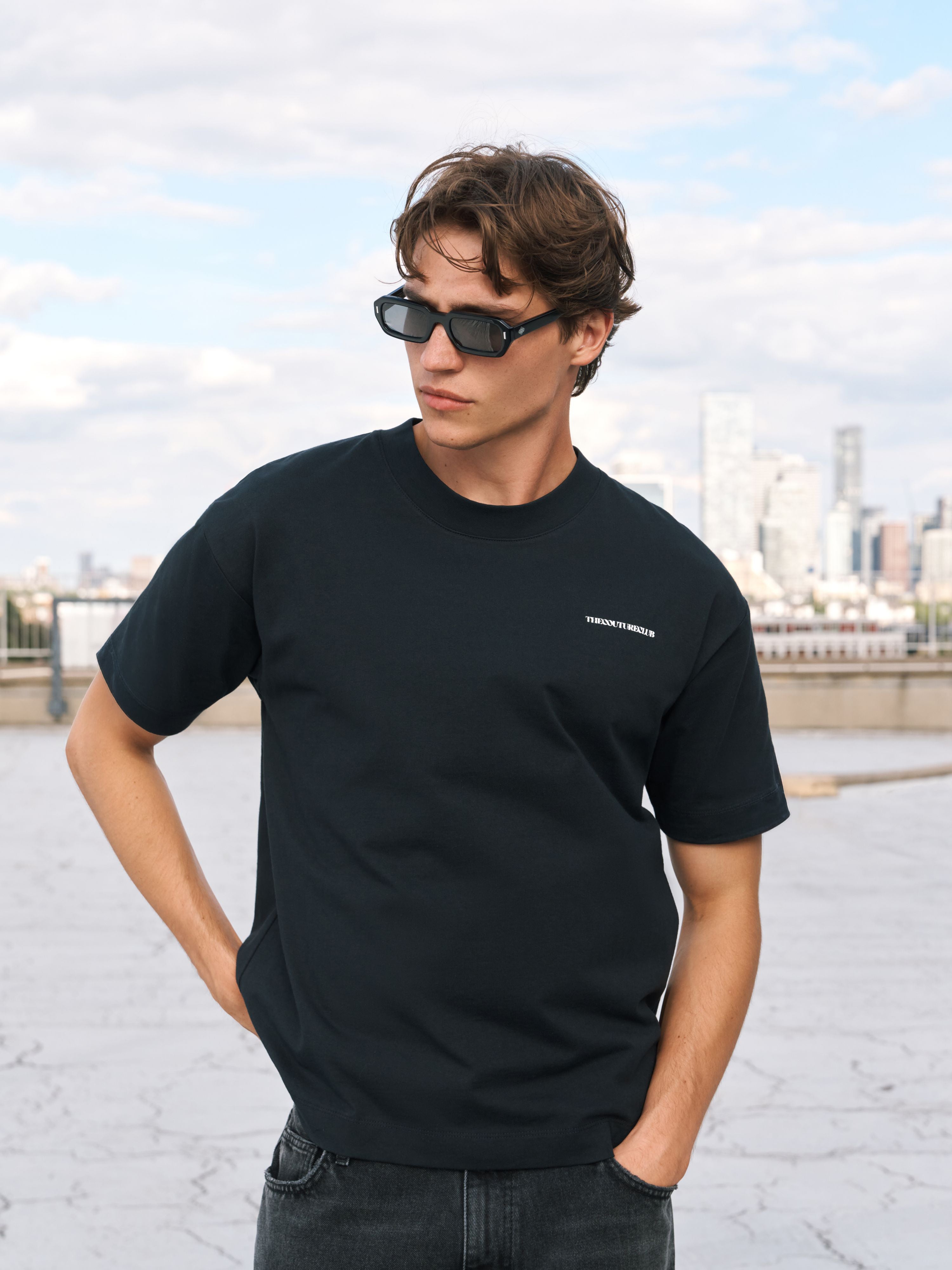 A man wearing a black short-sleeve crewneck t-shirt with small white text on the left chest and white graphic prints on both sleeves, paired with dark sunglasses and dark jeans, standing outdoors with a city skyline in the background.