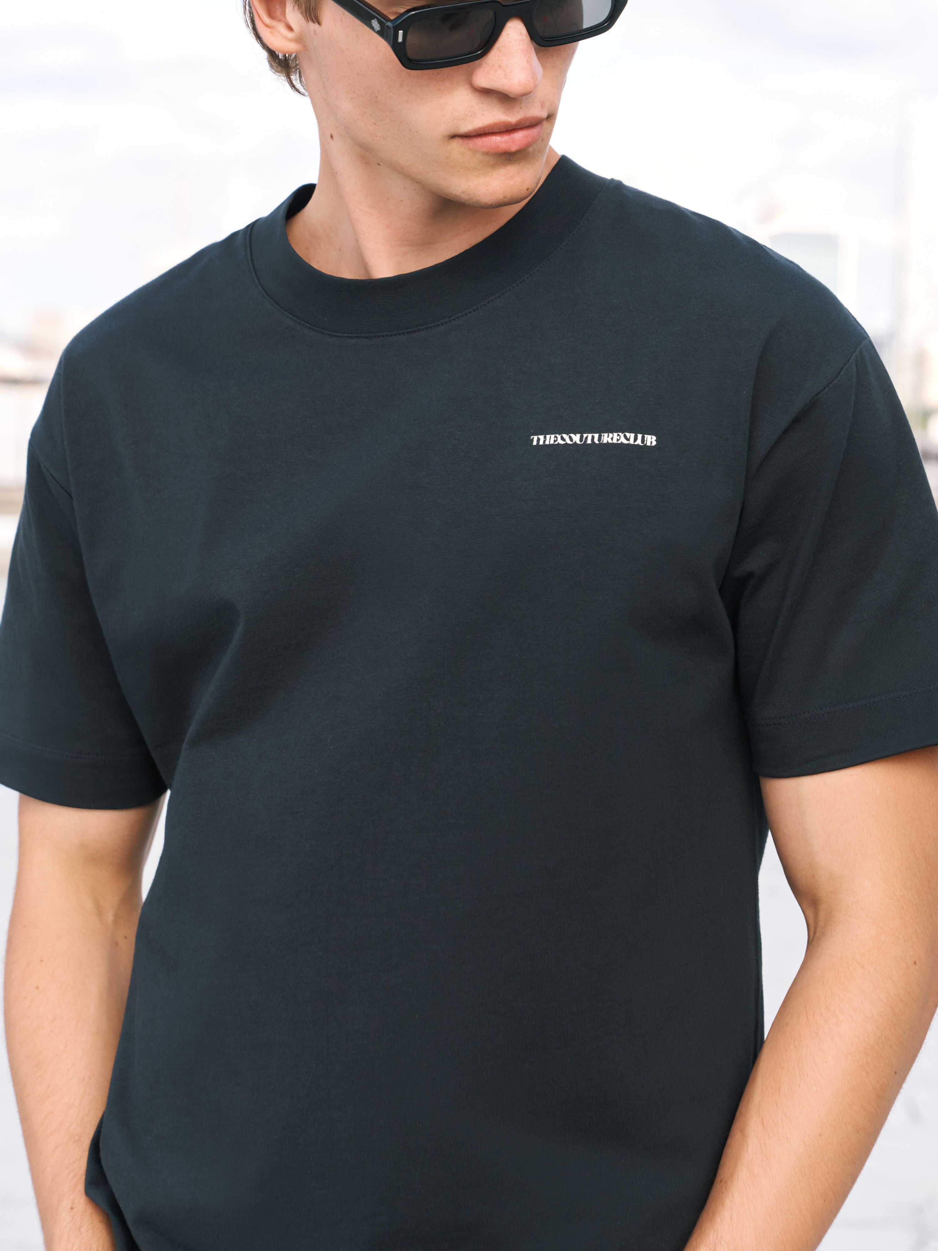 A man wearing a black crew-neck t-shirt with a small white 'The Couture Club' logo on the left chest, paired with black sunglasses.