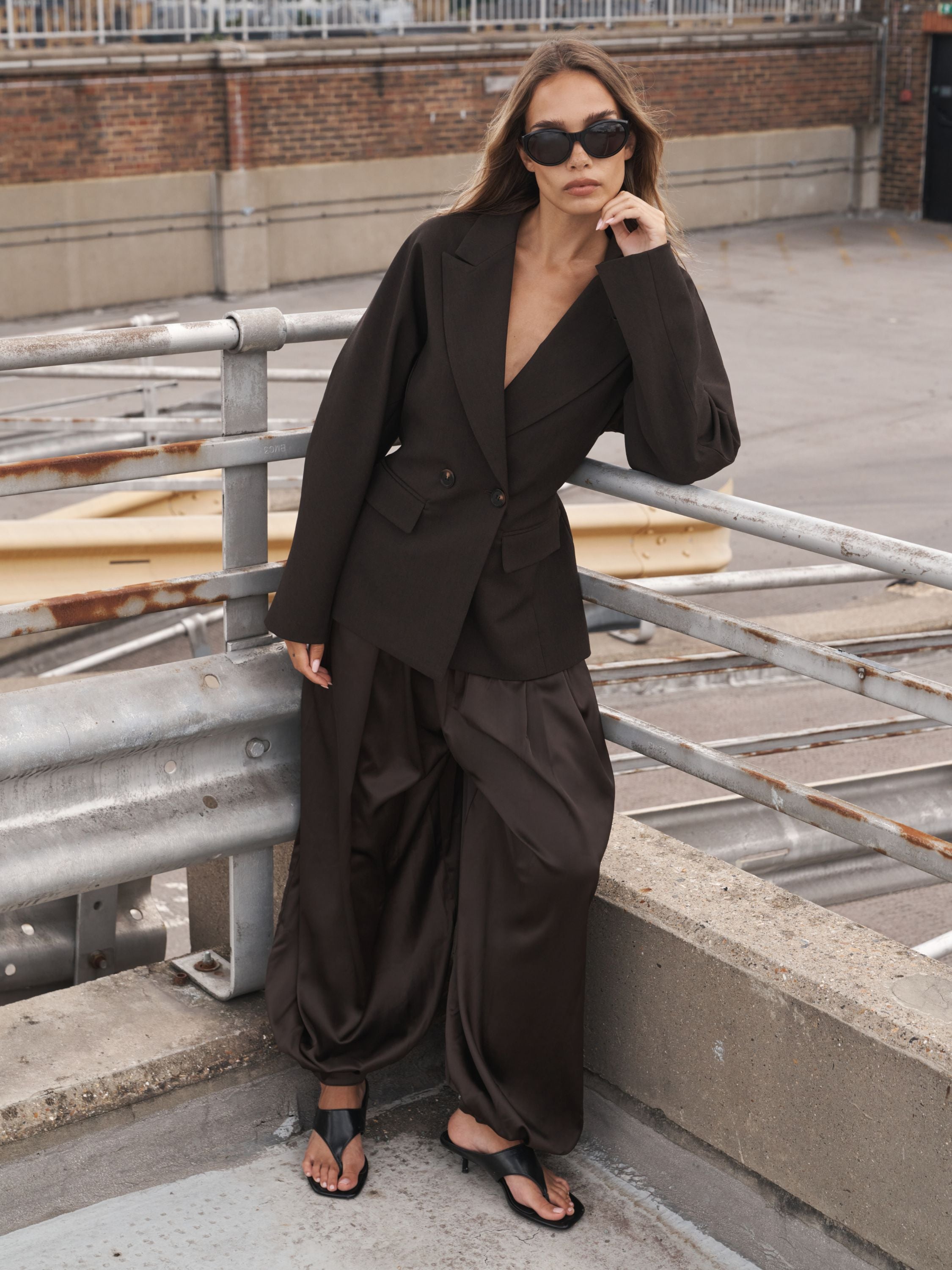 A woman wearing a dark brown tailored suit with a deep V-neck blazer and matching wide-leg pants, styled with black sunglasses and black sandals, posing outdoors on a bridge.