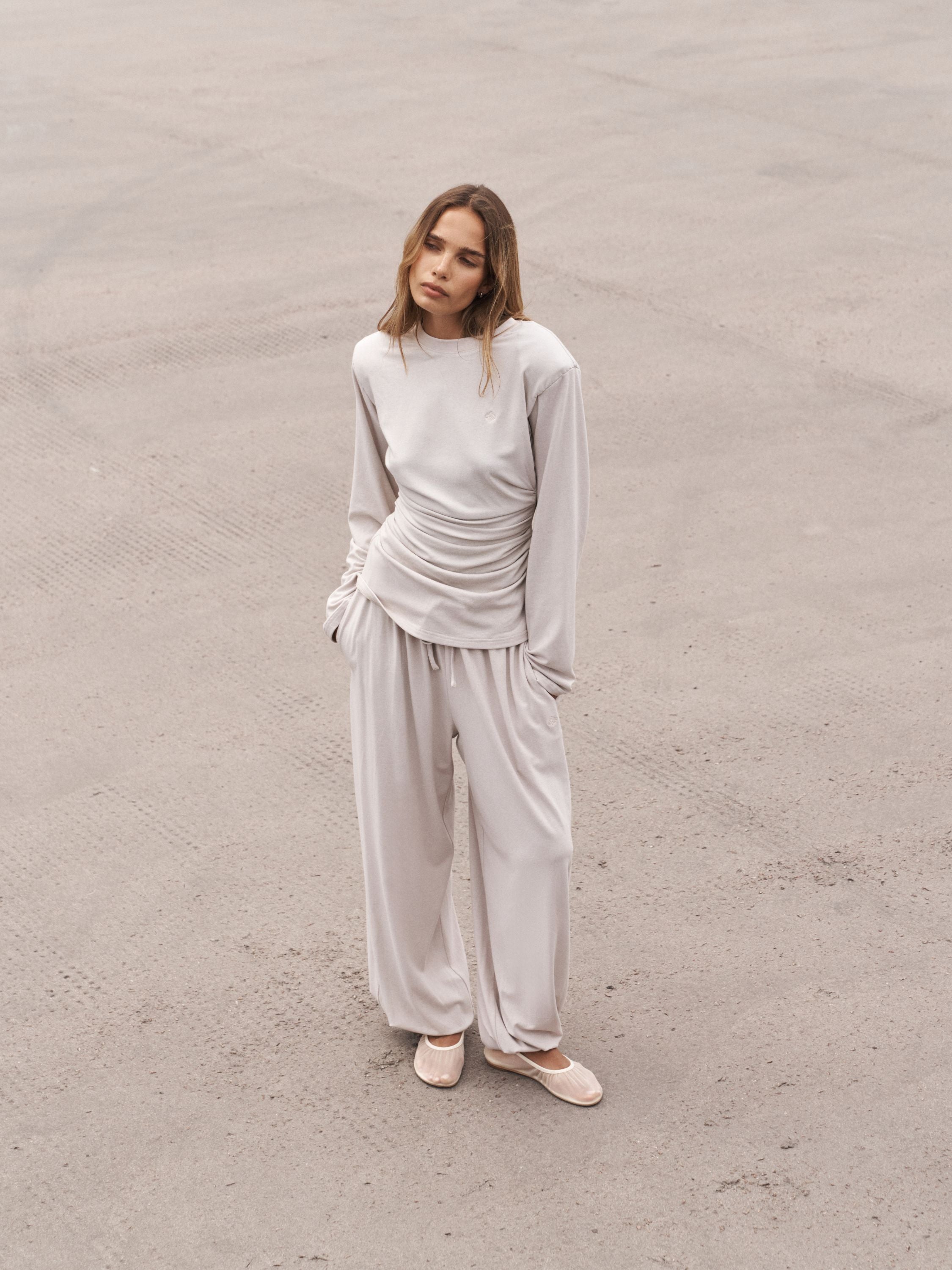 A woman stands on a concrete surface wearing a light beige long-sleeve top and matching wide-leg pants, paired with beige flats.