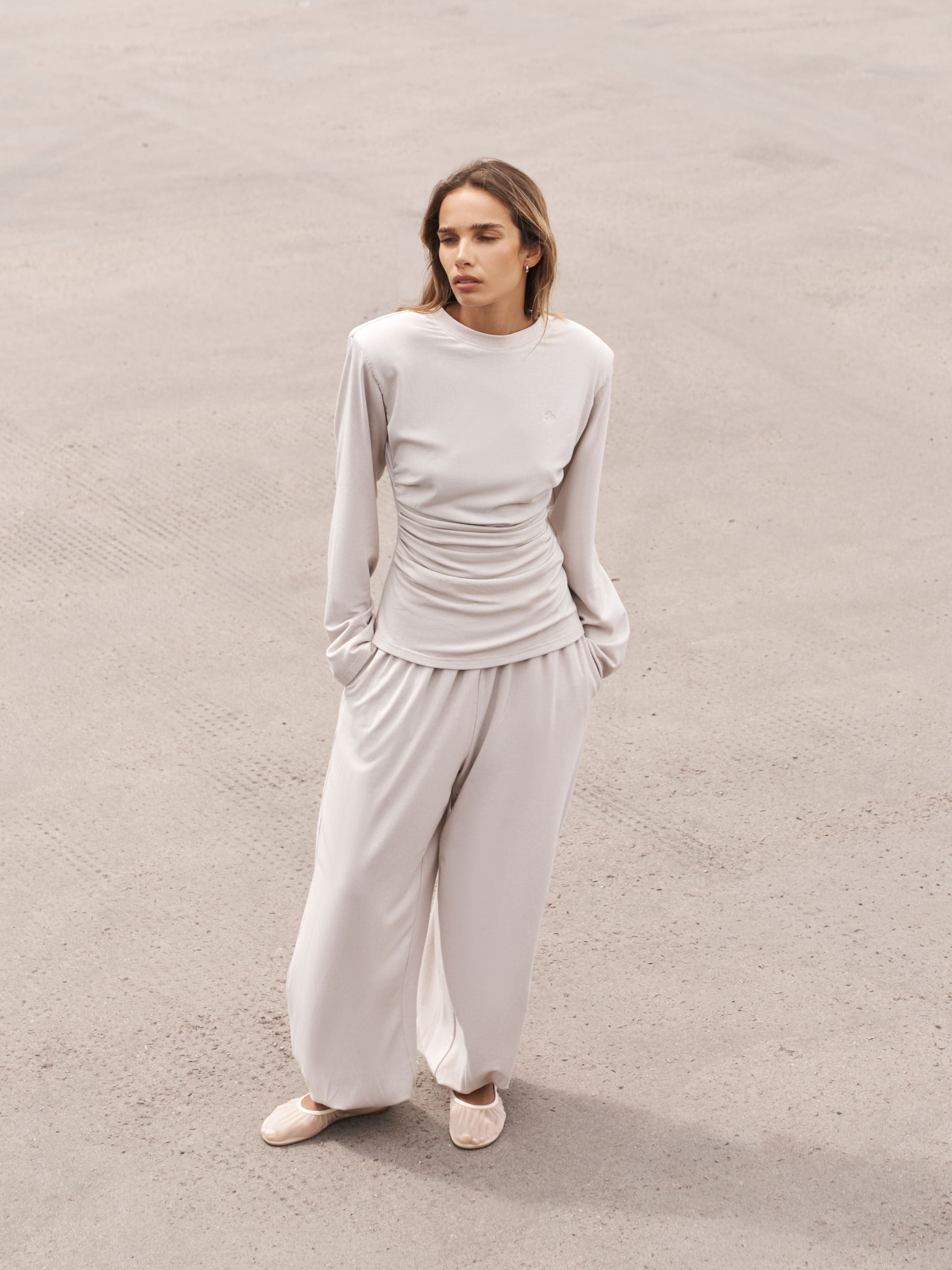 A woman stands on a textured concrete surface wearing a light beige ribbed knit two-piece set consisting of a long-sleeve top with a ruched waist and matching wide-leg pants.