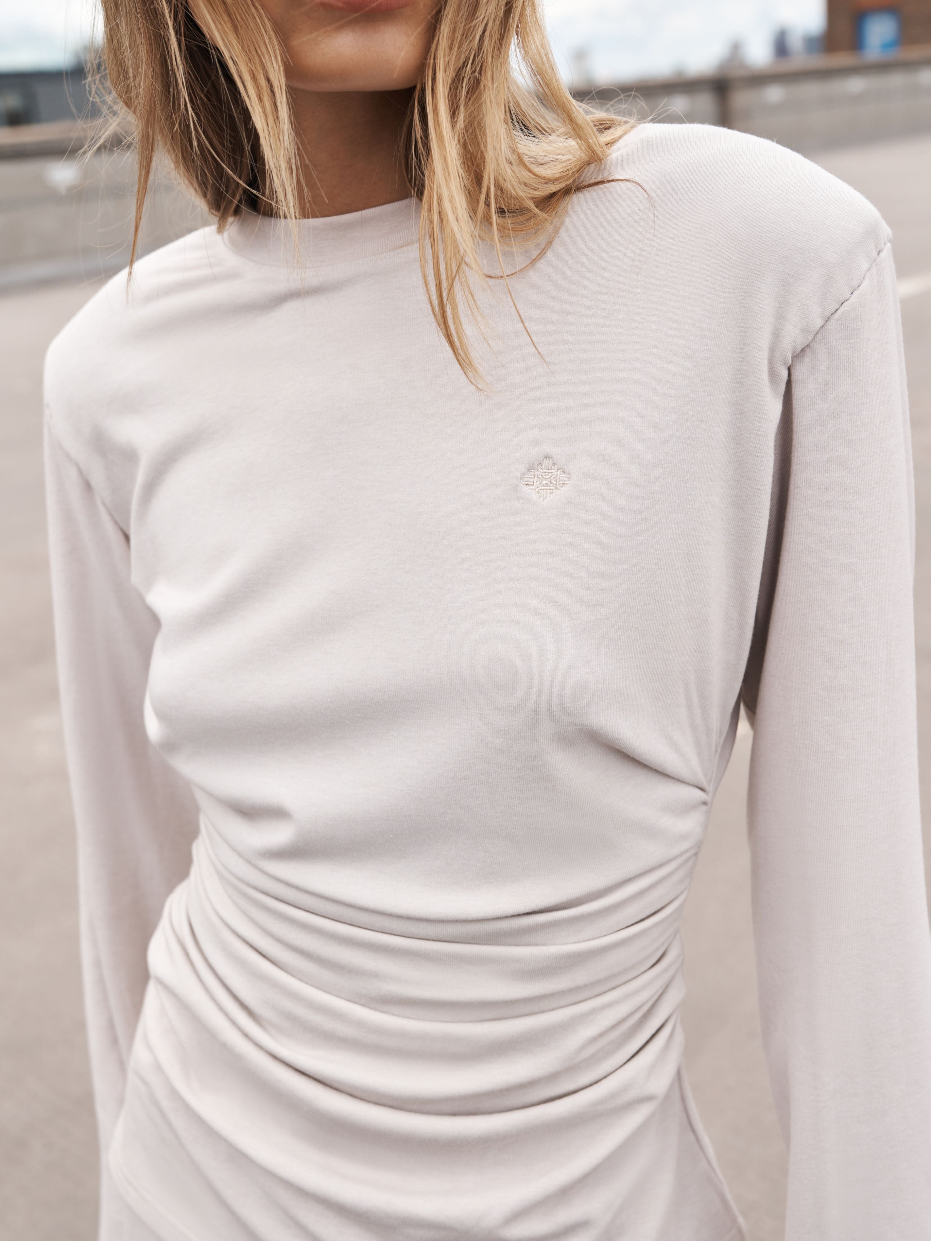 A close-up of a model wearing a light beige long-sleeve top with a gathered ruched waist and a small tonal diamond logo on the chest, set against an outdoor blurred background.