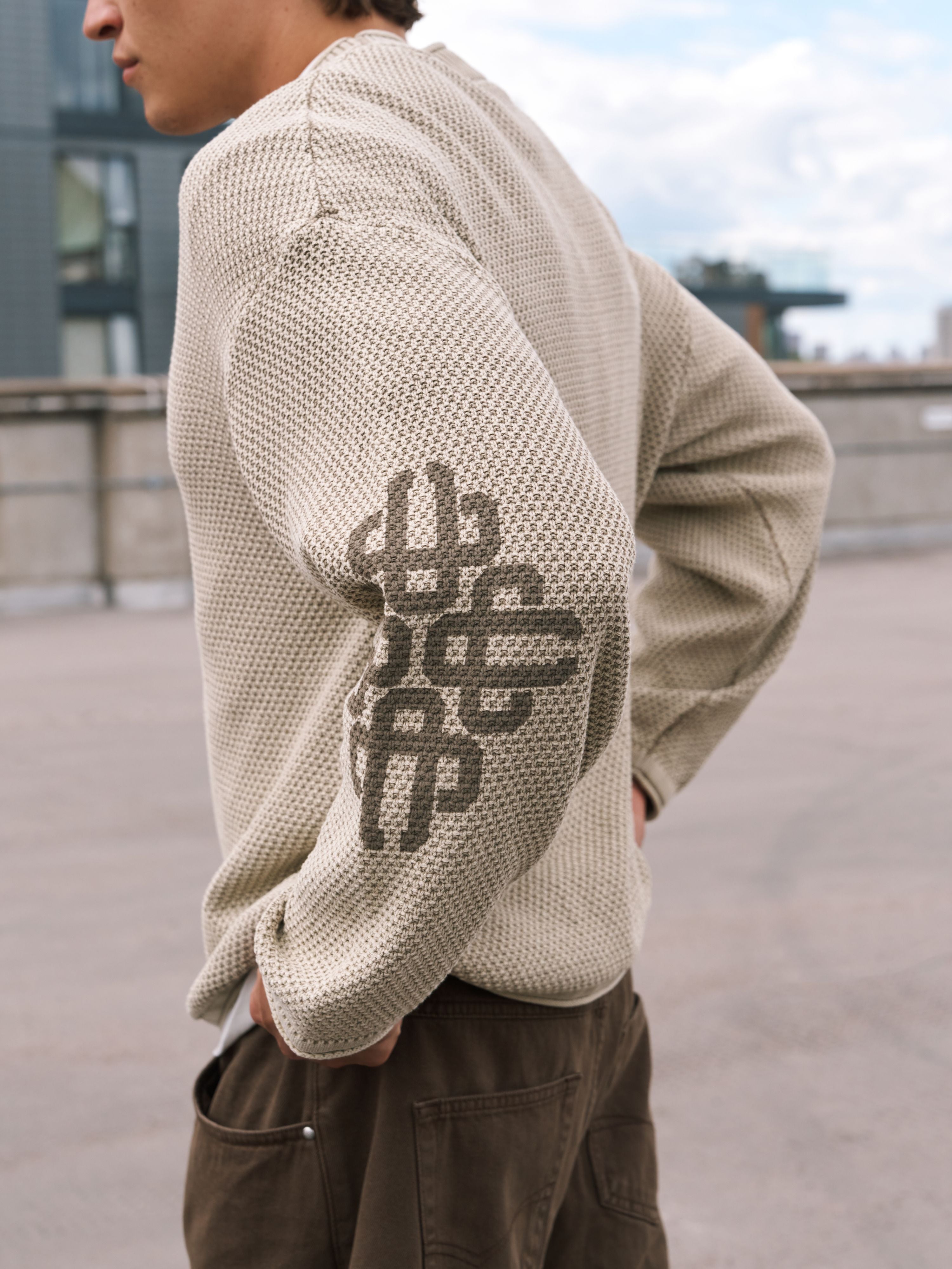 A person wearing a beige textured knit sweater with a dark gray woven geometric logo on the upper sleeve, paired with brown pants, photographed outdoors on a rooftop.