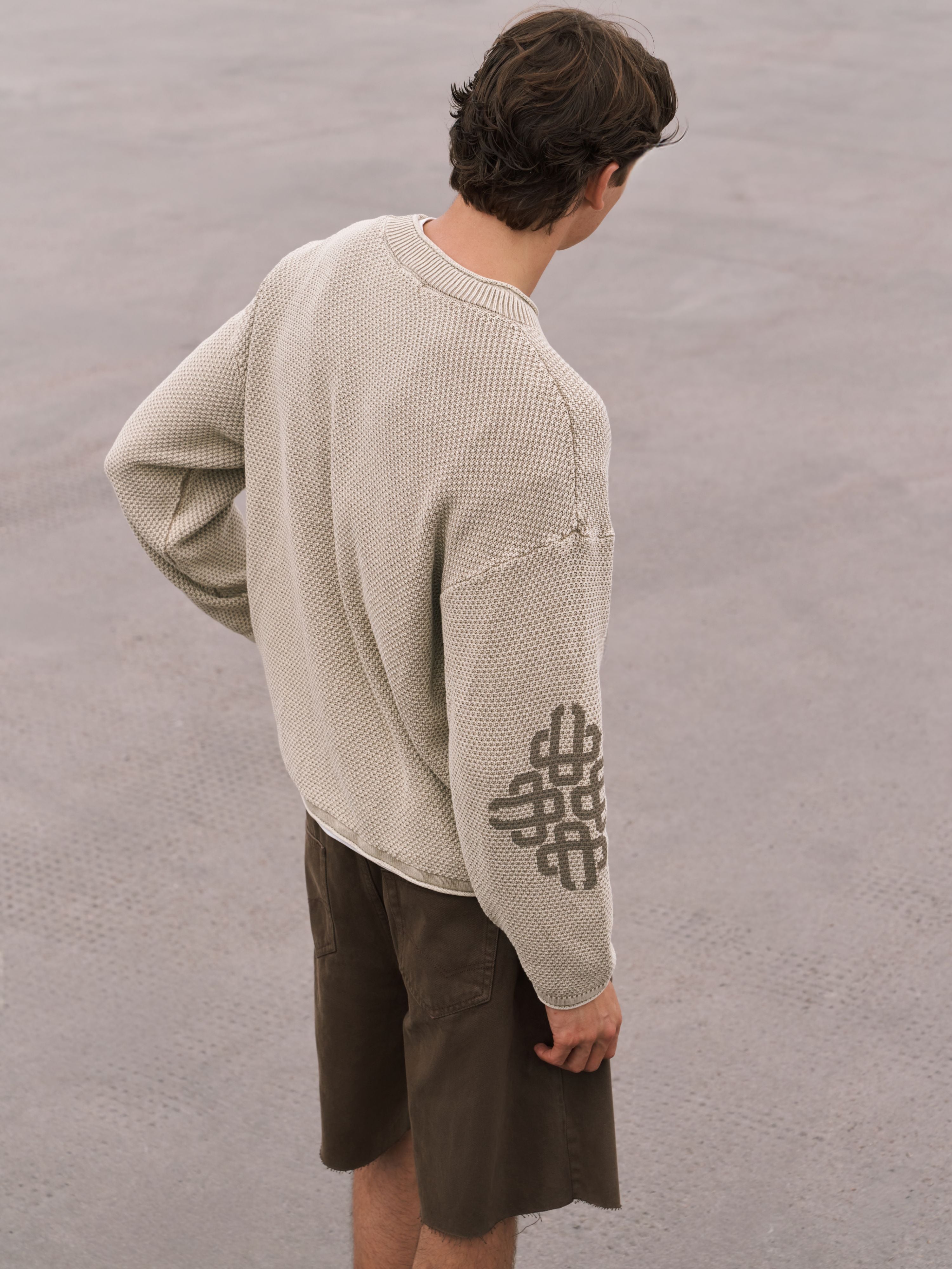 A man wearing a beige textured knit sweater with a dark gray geometric pattern on the sleeve, paired with brown shorts, photographed from behind against a neutral gray concrete background.