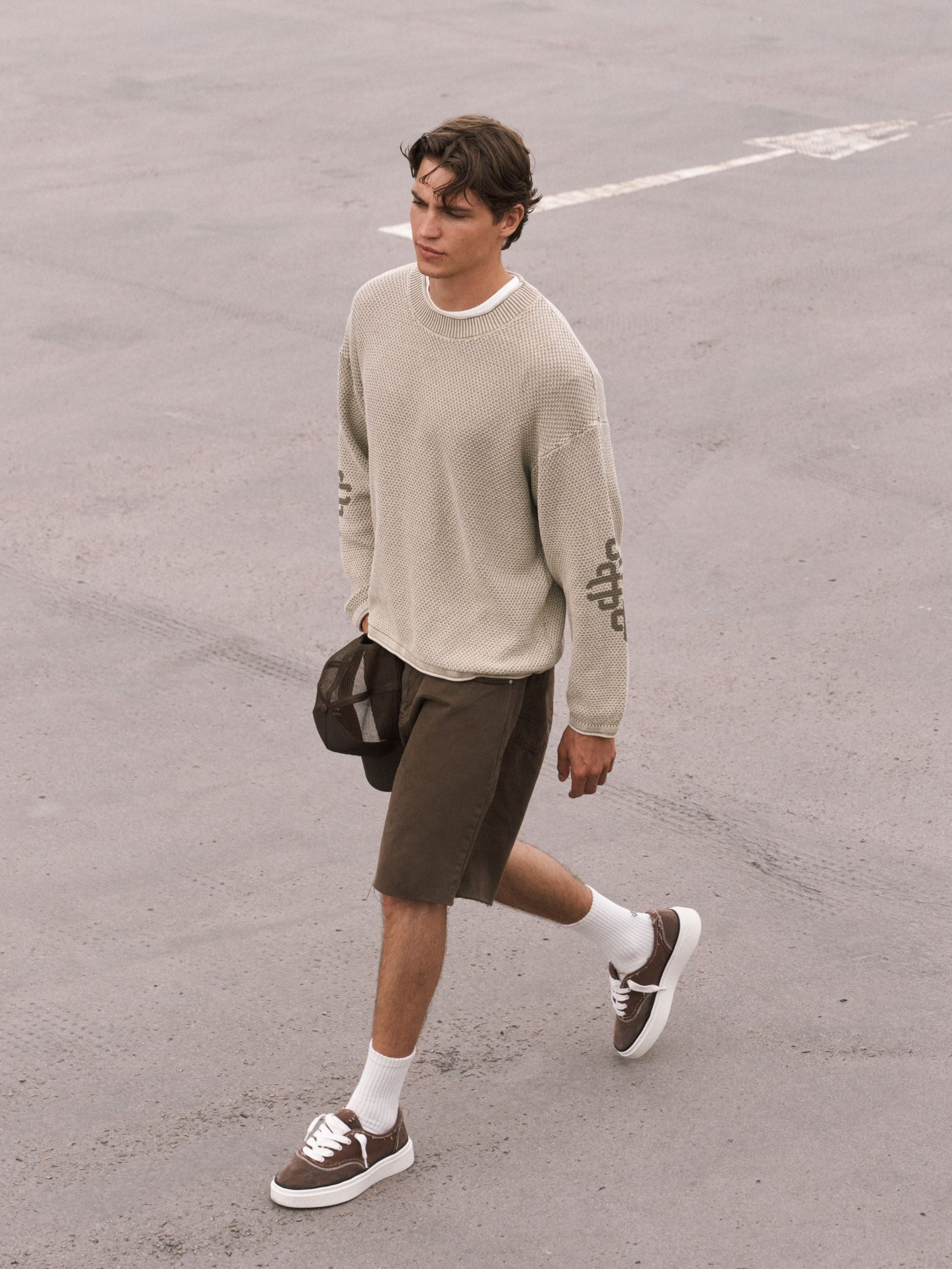 A man walking on a gray paved surface wearing a light beige crewneck sweater, dark brown shorts, white socks, and brown boat-style sneakers, with a dark cap in his hand.