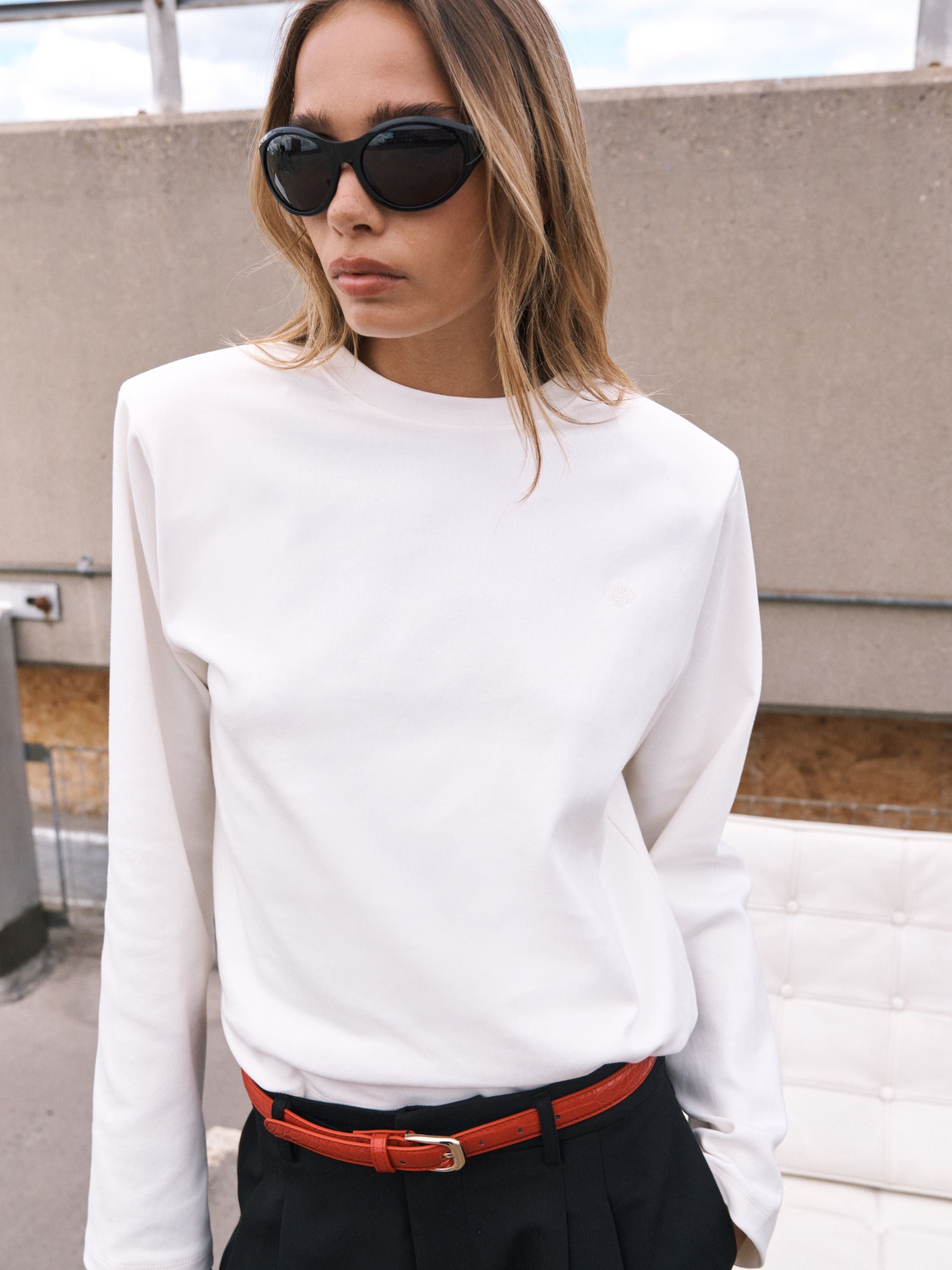 A woman wearing a white long-sleeve crewneck top, black sunglasses, and a red belt, standing outdoors against a concrete wall.