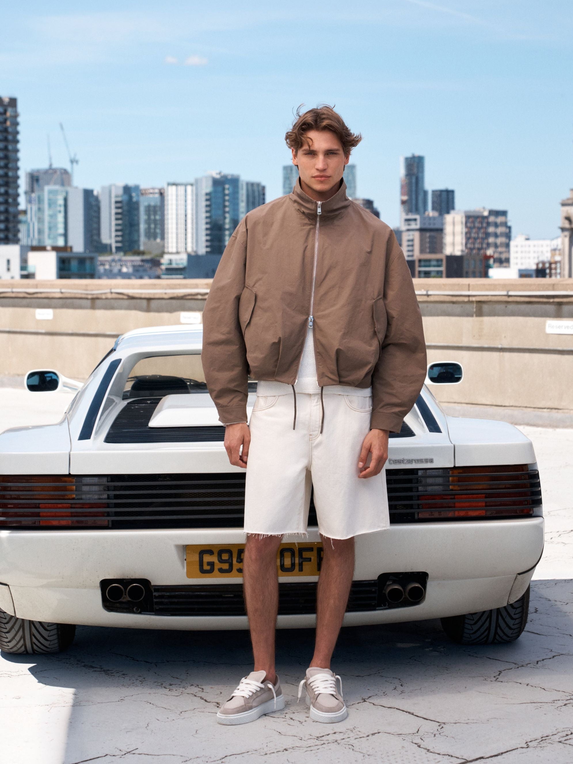 A man stands in front of a white classic sports car on a rooftop, wearing a brown zip-up jacket, white shorts, and white sneakers. The city skyline is visible in the background under a blue sky.