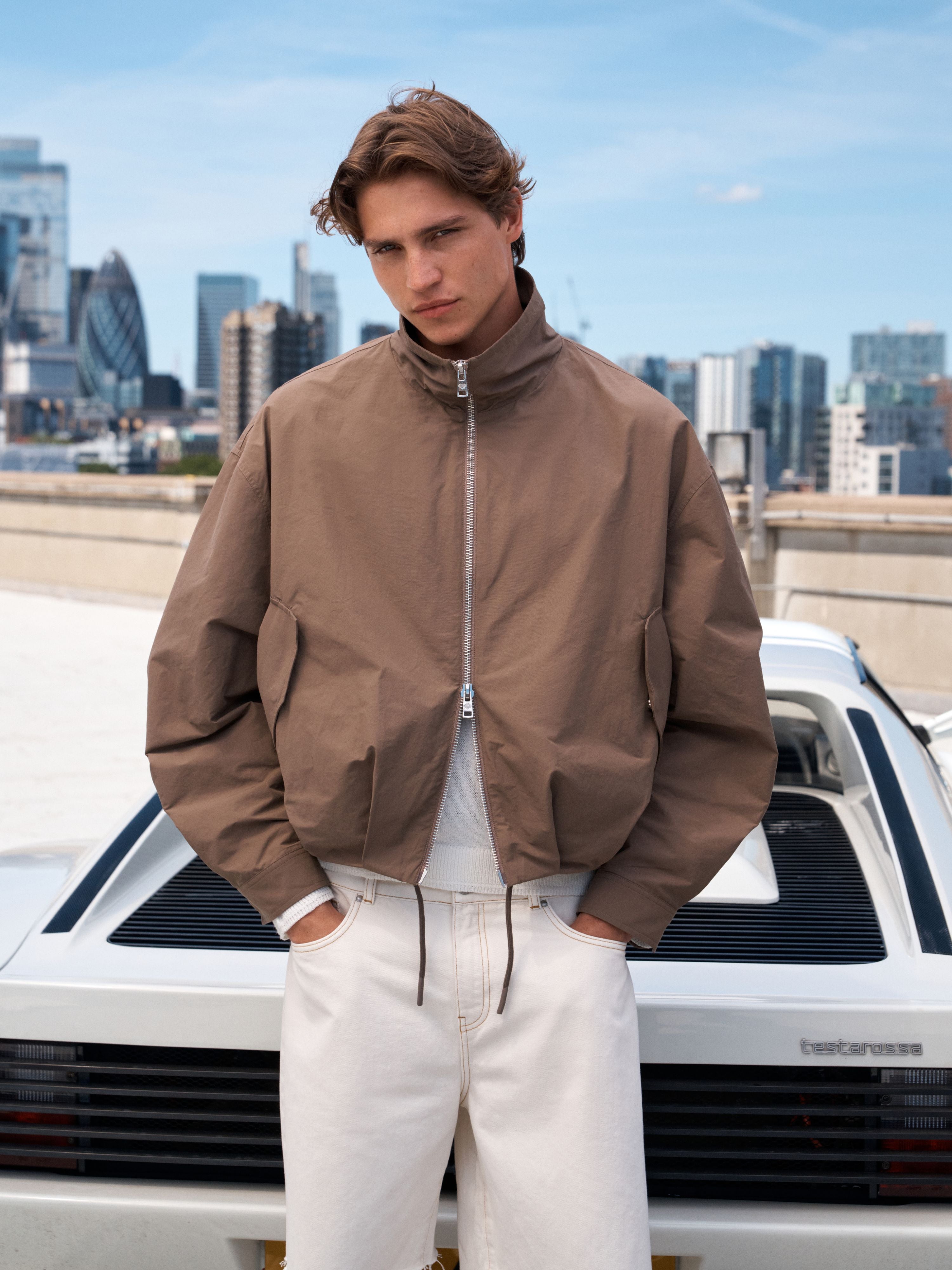 A man wearing a brown zip-up jacket, white sweatshirt, and white drawstring pants stands in front of a white classic car with a city skyline in the background.