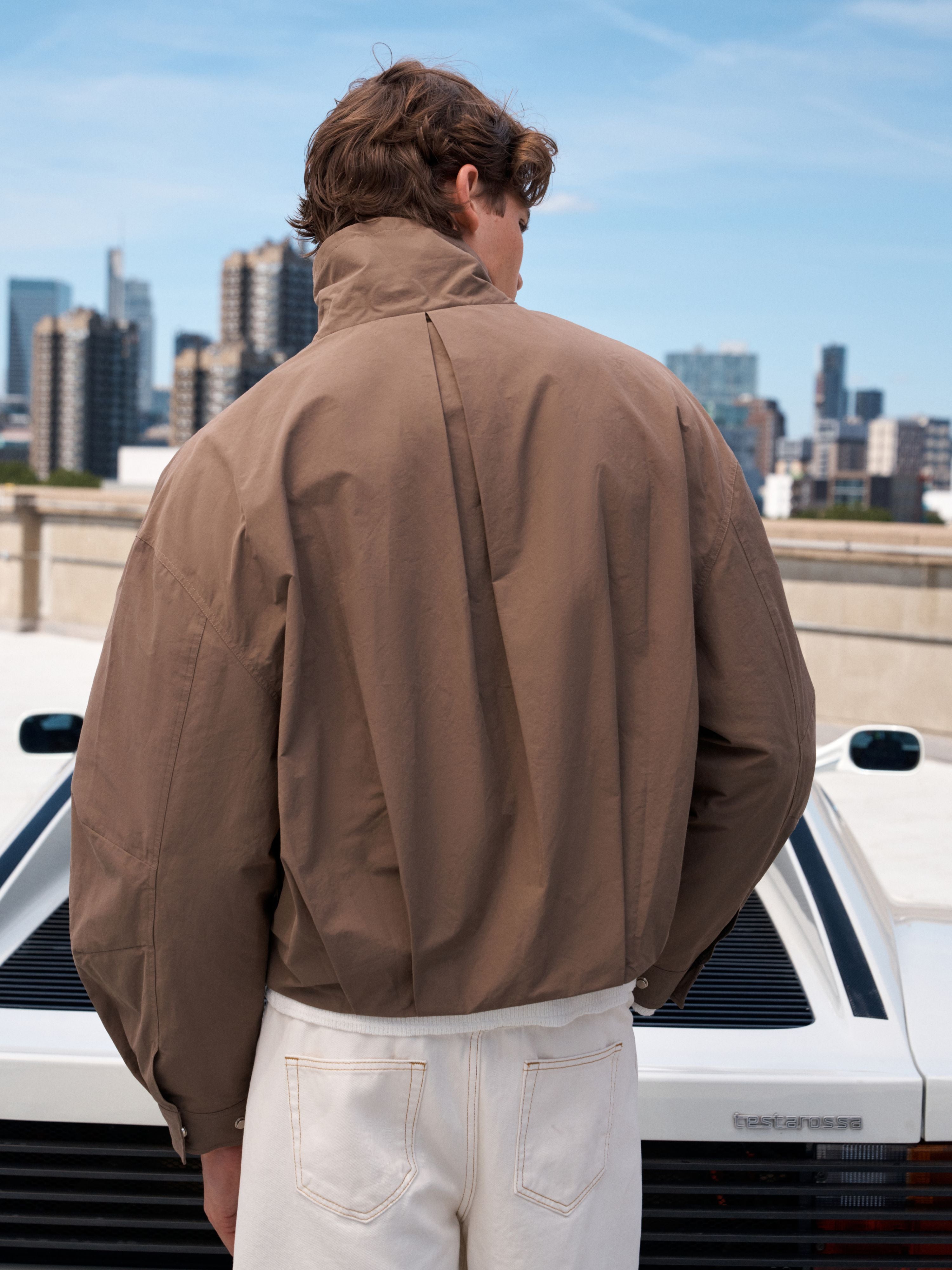 A man viewed from behind wearing a brown lightweight jacket and off-white pants, standing outdoors in front of a white car with a city skyline in the background.