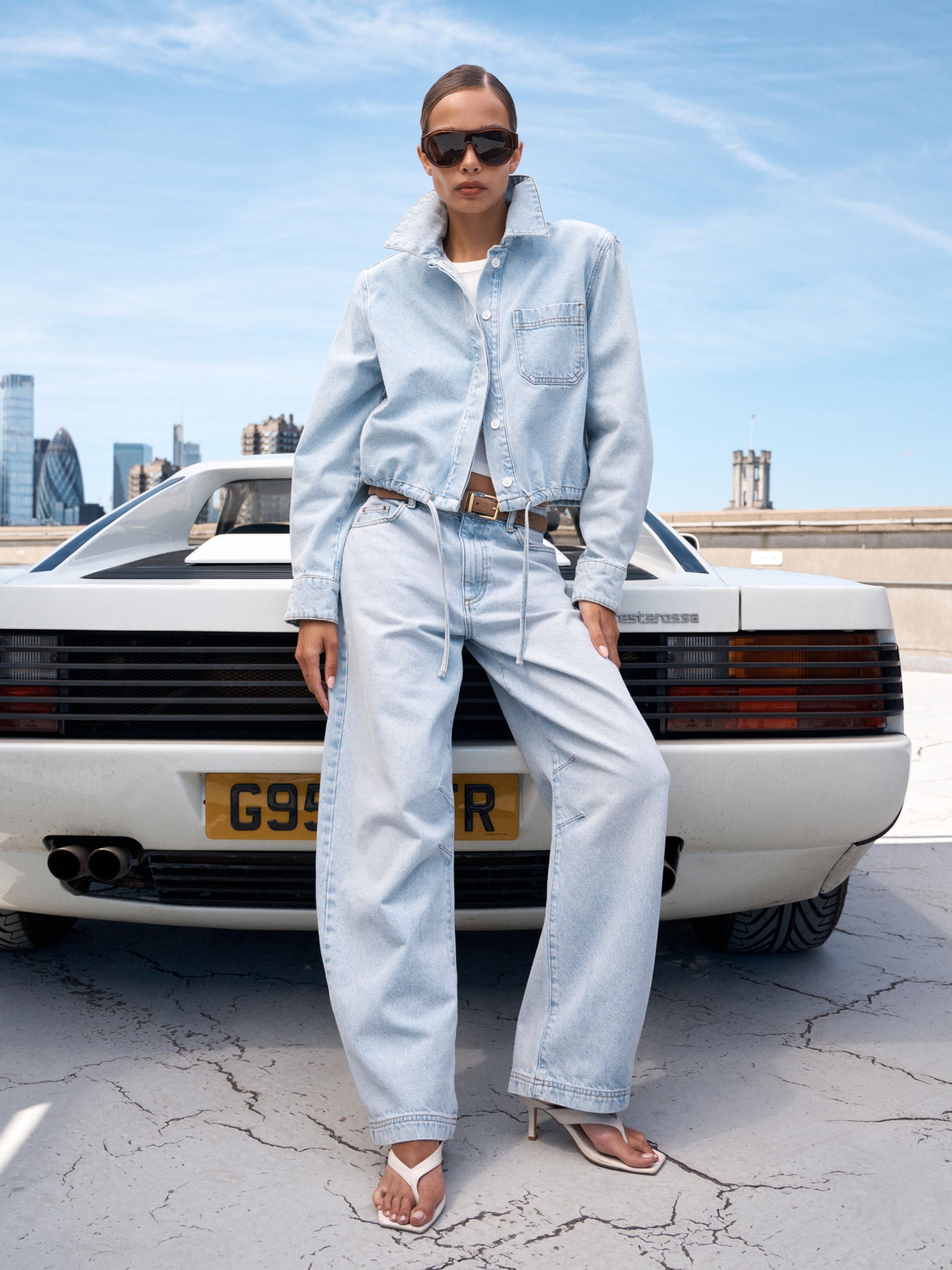 A model stands in front of a white classic car on a rooftop, wearing a light-wash denim button-up shirt and matching wide-leg jeans with a brown belt and white sandals.