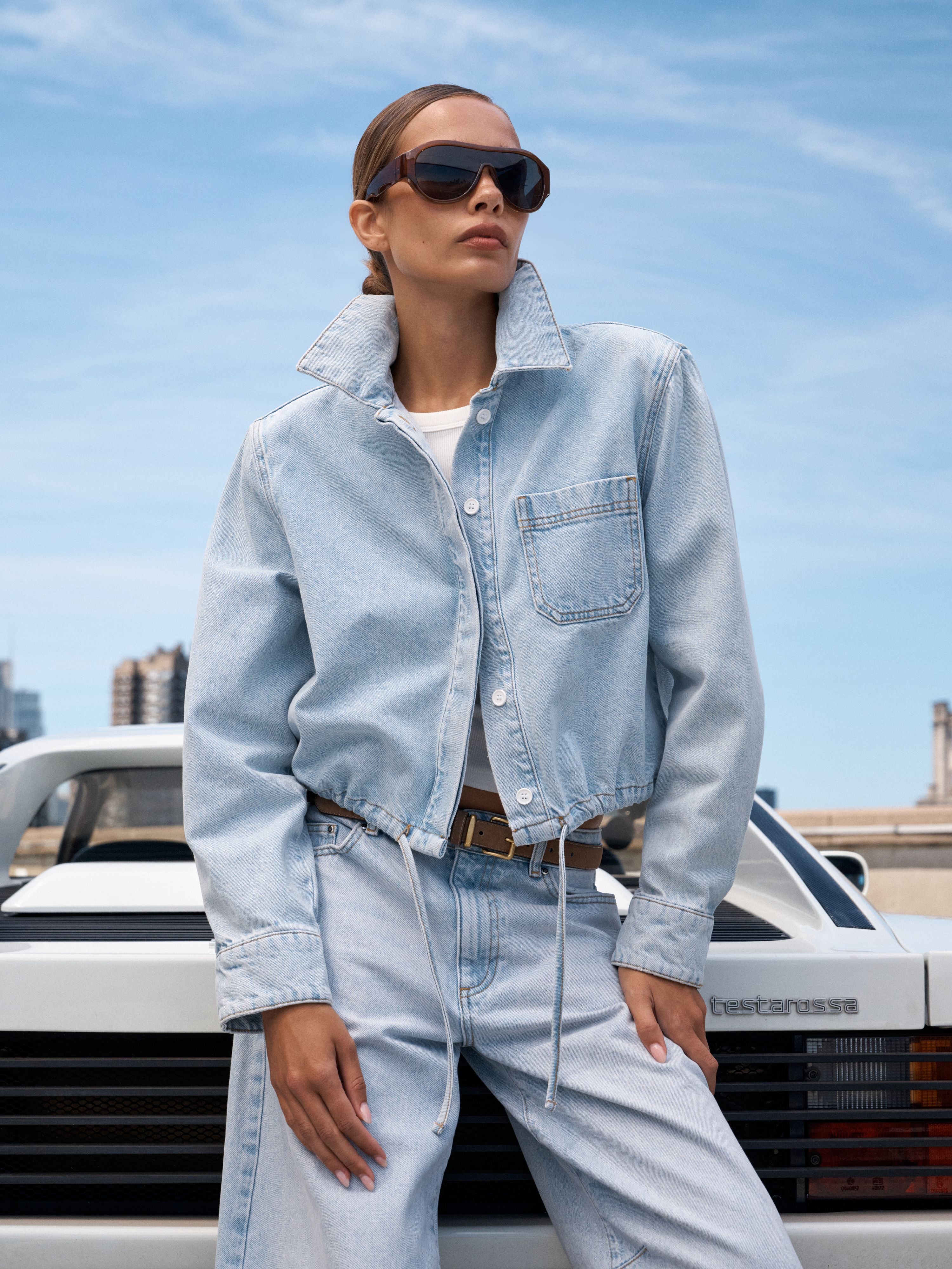 A woman wearing a light-wash denim jacket and matching wide-leg jeans stands outdoors in front of a white car under a blue sky.