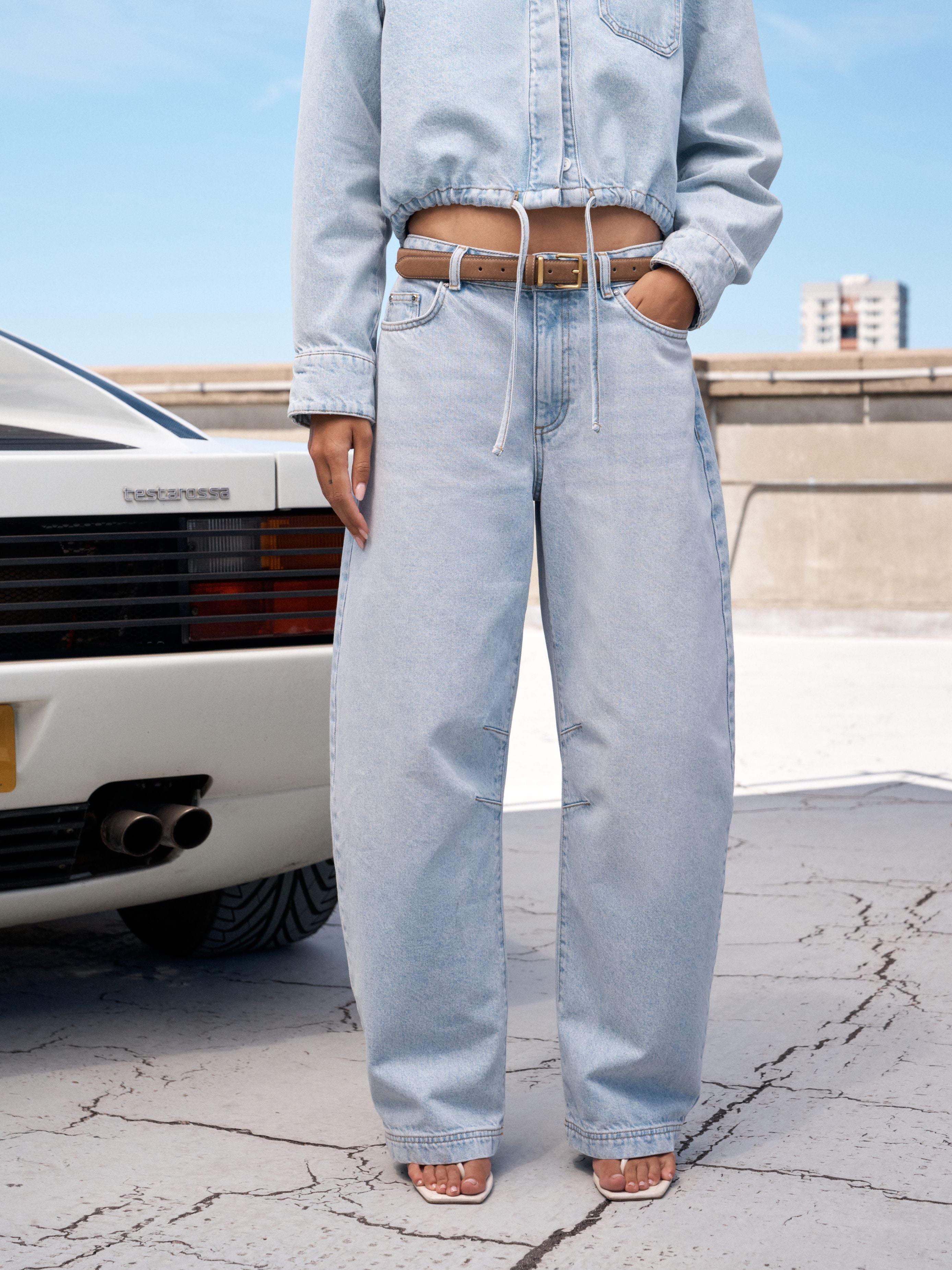 A person wearing light-wash wide-leg denim jeans with a drawstring waist, paired with a cropped light denim shirt and tan sandals, standing outdoors next to a white car on a cracked concrete surface.