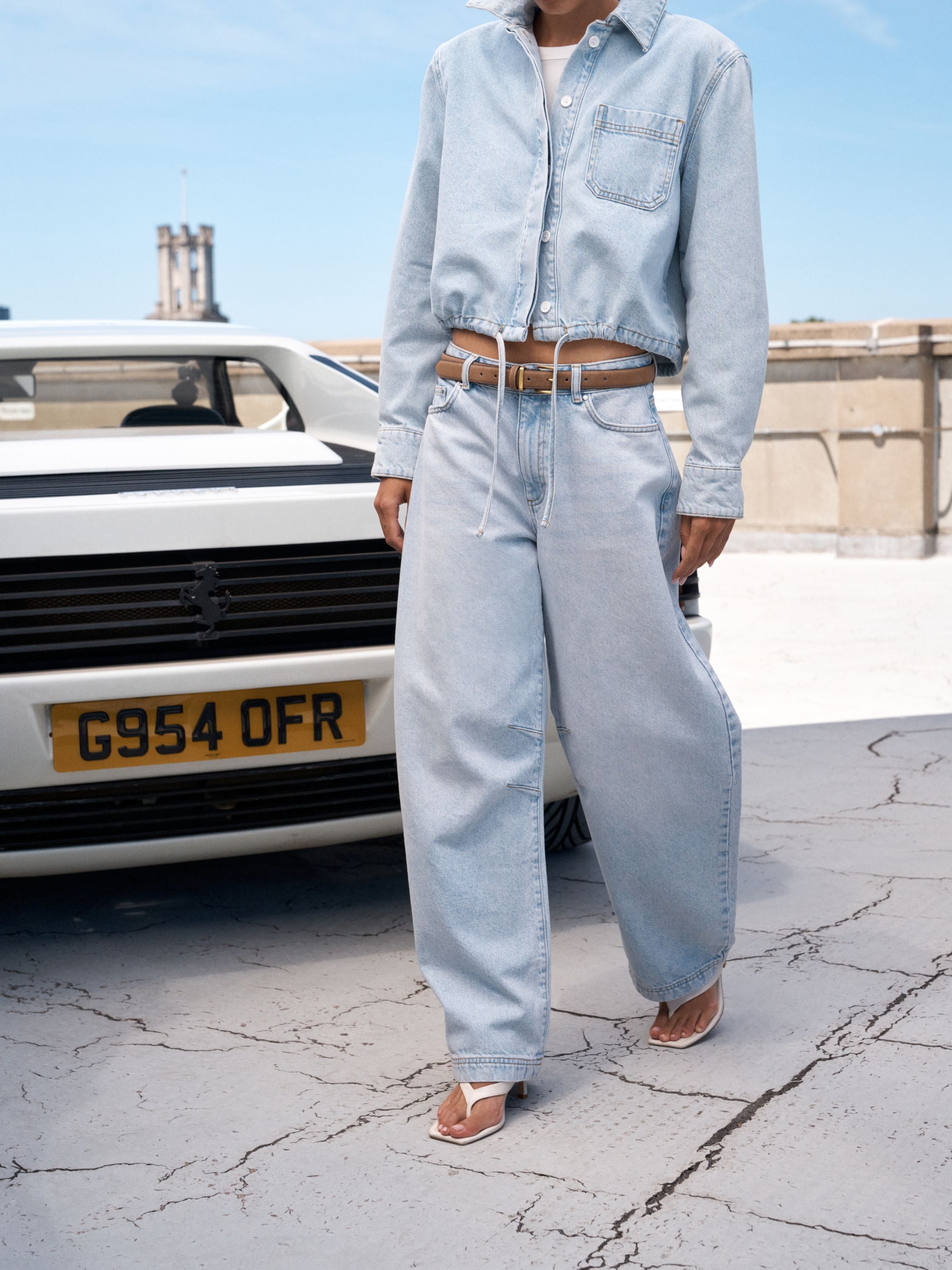 Woman wearing light blue barrel leg jeans next to a white classic car.