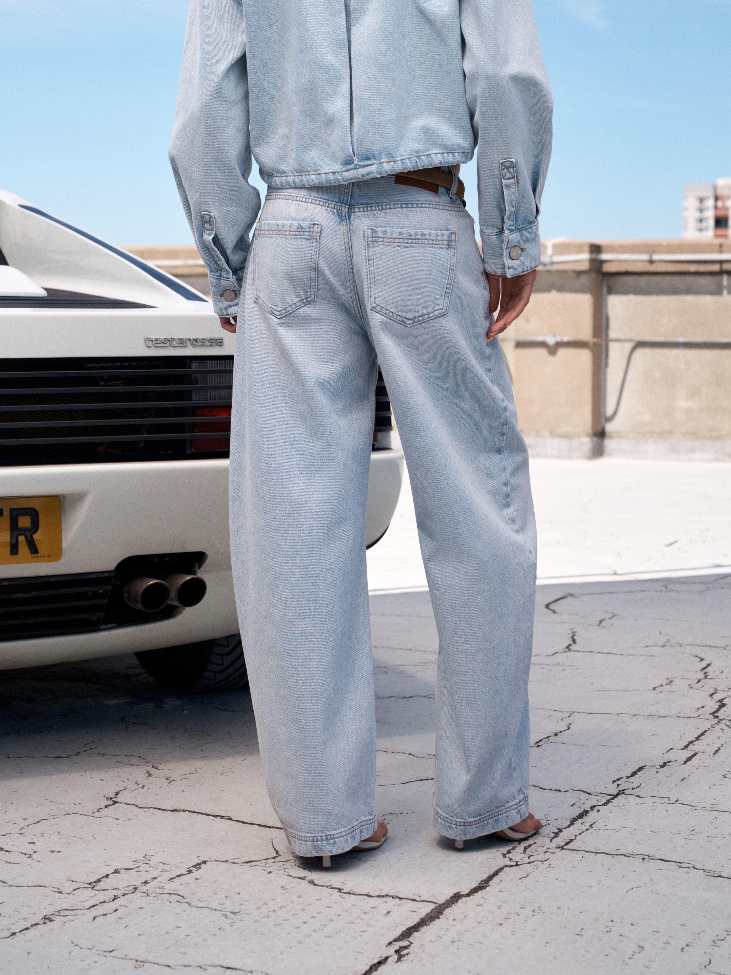 A person viewed from behind wearing very light blue wide-leg denim jeans, a matching light blue cropped jacket, and brown sandals, standing outdoors beside a white car on a cracked concrete surface.