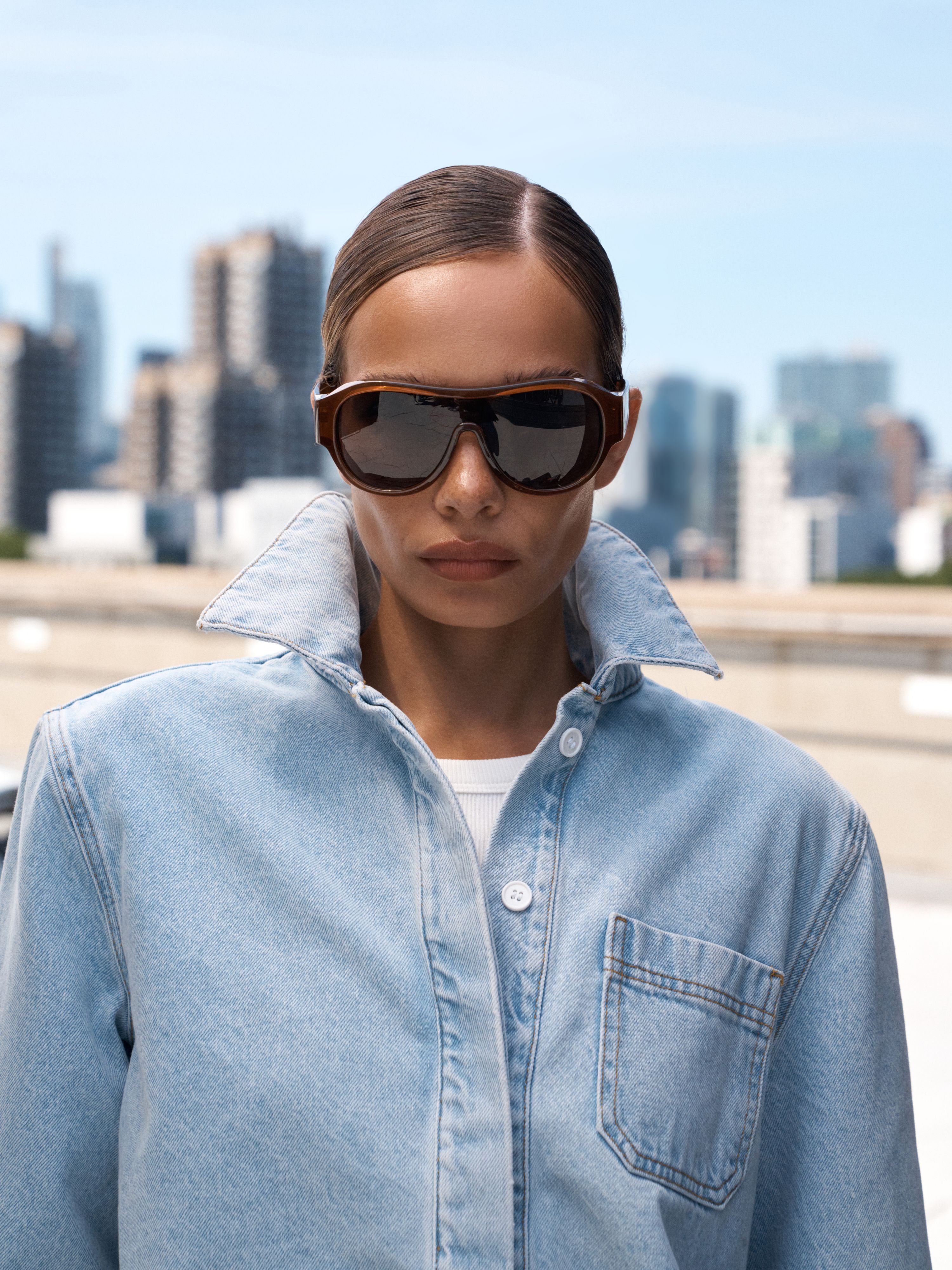 A model wearing oversized dark sunglasses with brown frames and a light blue denim shirt, set against a blurred city skyline background.