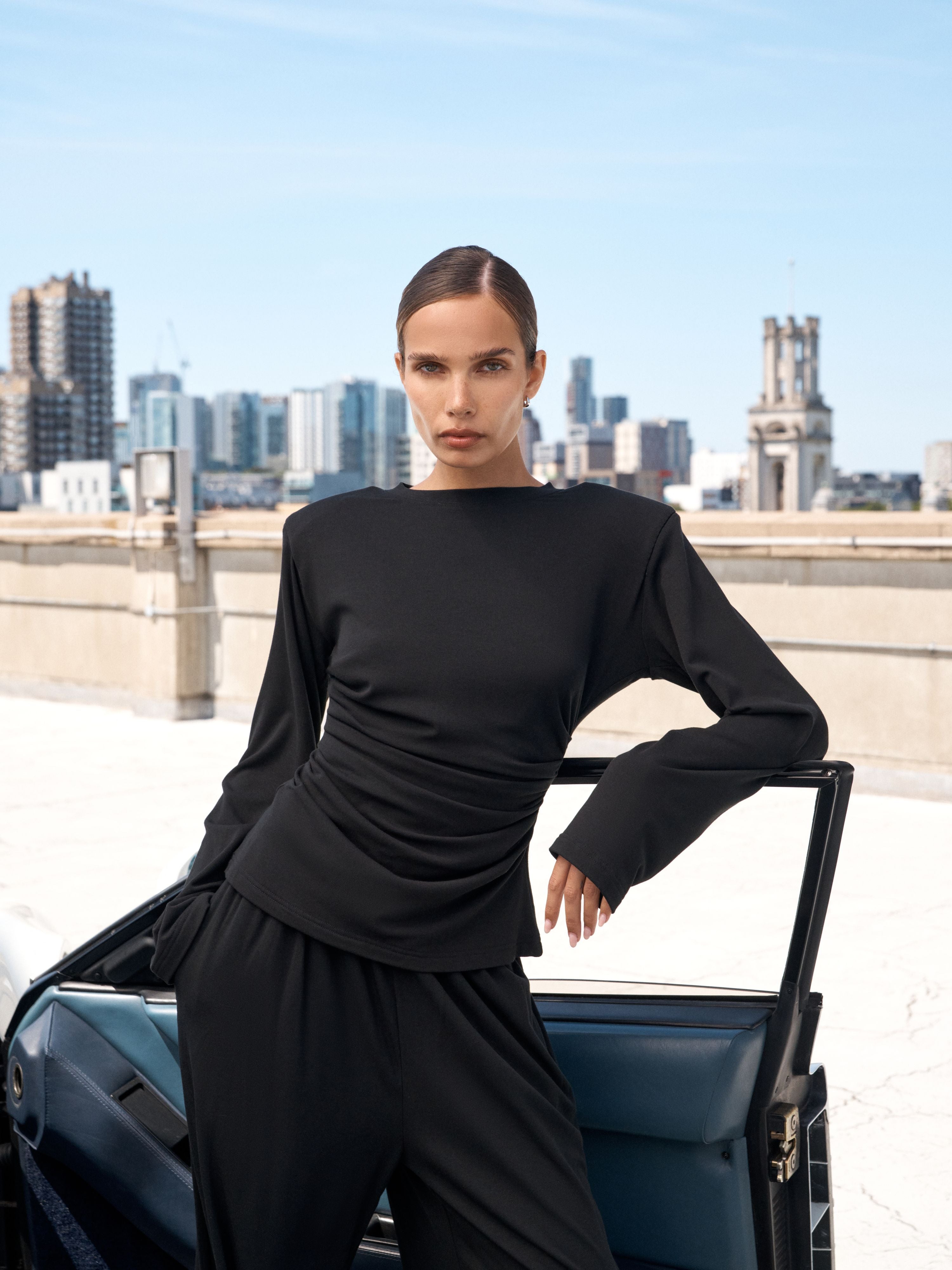 A model wearing a black draped long-sleeve top and matching wide-leg trousers, leaning against an open blue car door on a rooftop with a city skyline in the background.