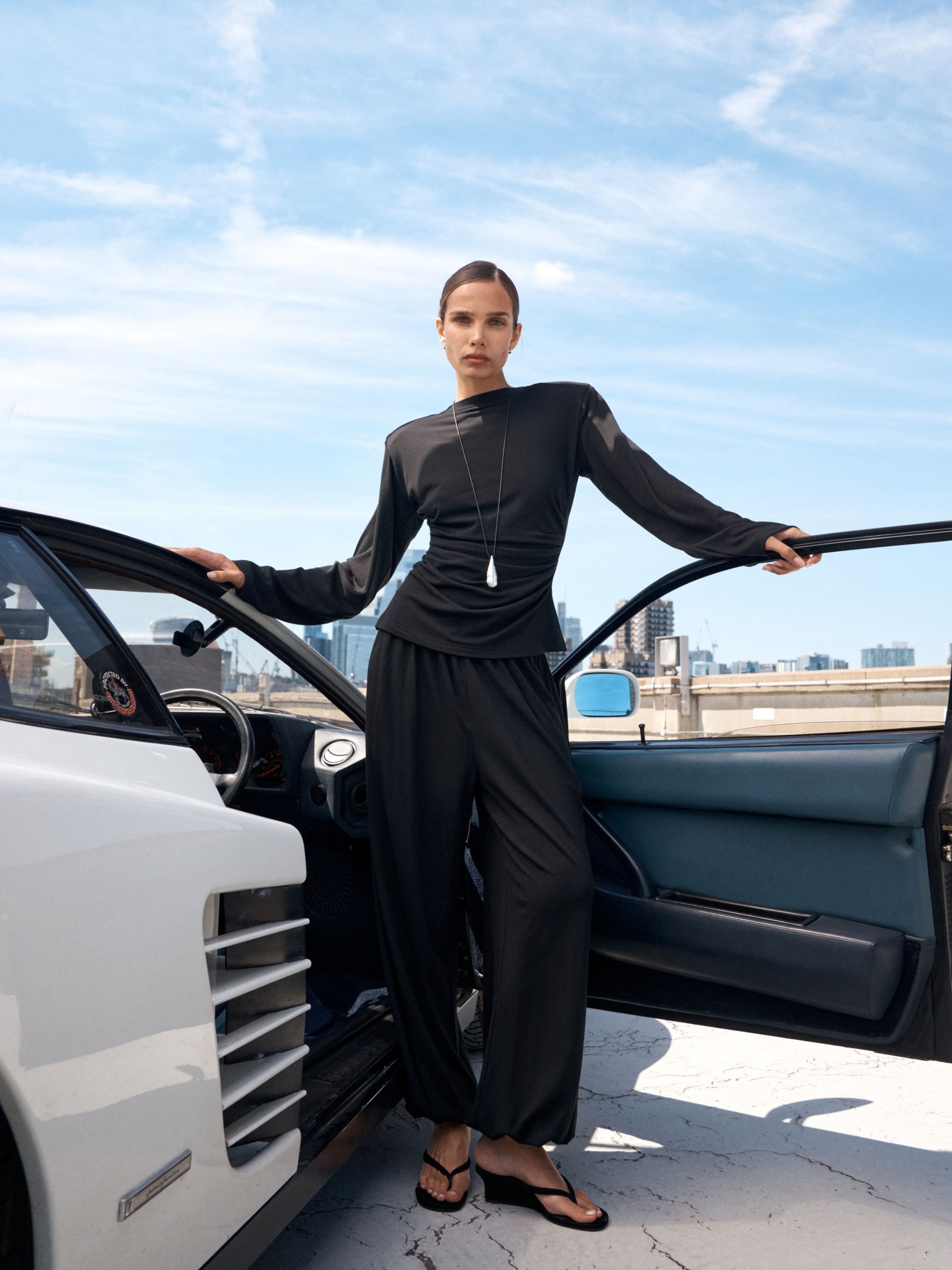 A model wearing a black long-sleeve top and matching wide-leg trousers stands beside an open white car on a rooftop terrace under a blue sky.