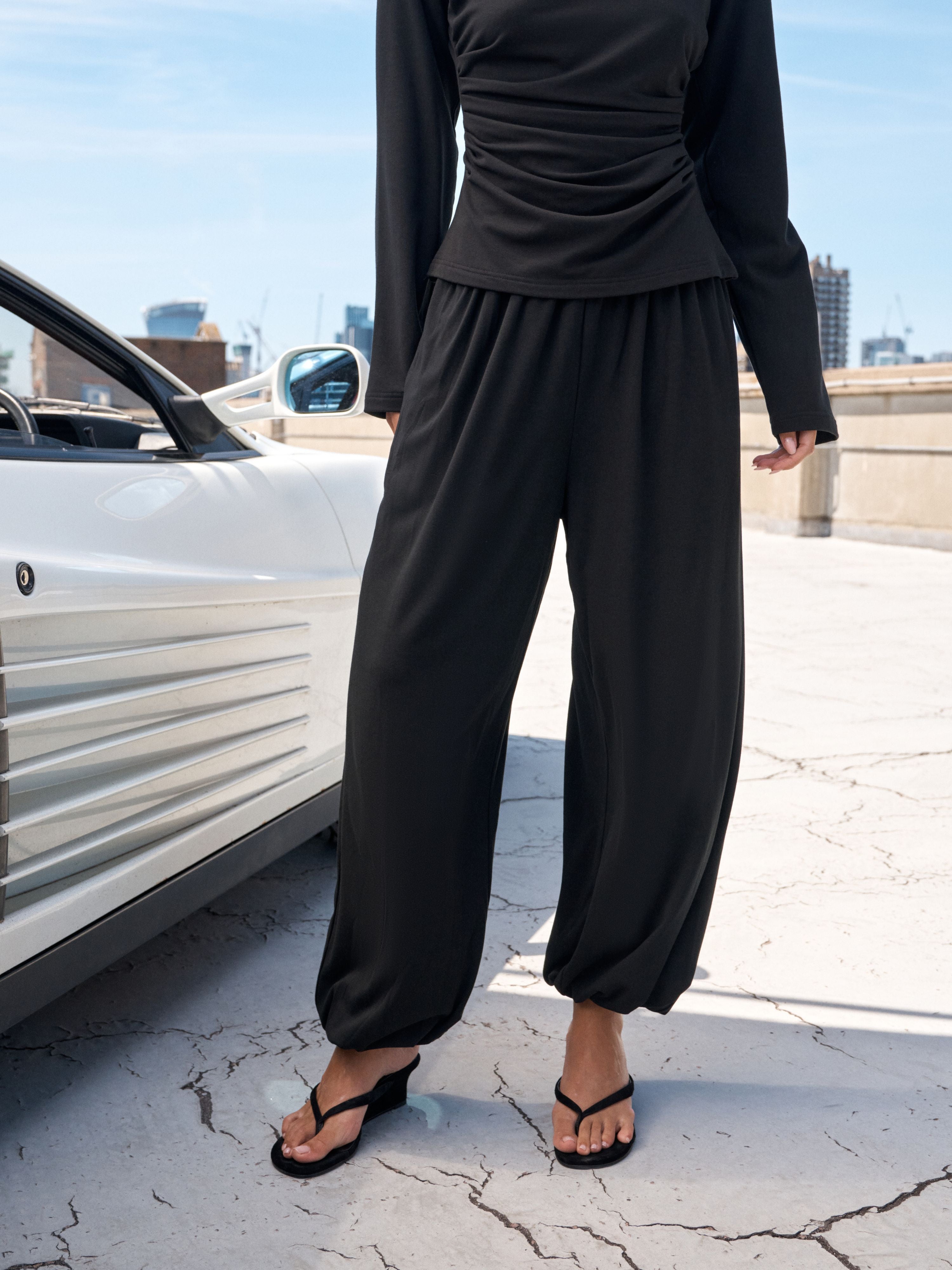 A person wearing a black long-sleeve top and matching loose black pants, standing outdoors beside a white car on a cracked concrete rooftop under a blue sky.
